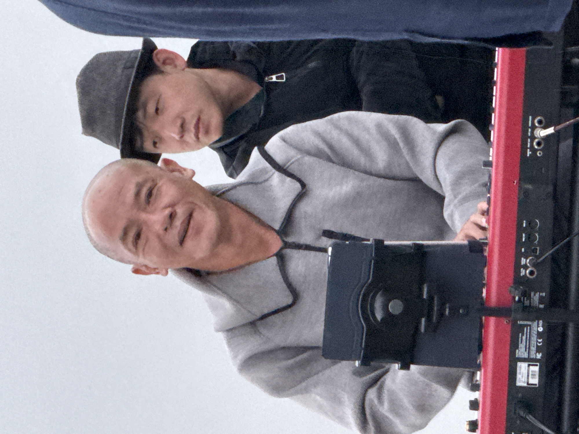 An outdoor, overcast medium shot shows three people standing behind a red keyboard. The person on the left, facing the camera, is a bald East Asian man in a gray hooded jacket. He is smiling and has his hands on the keyboard. A tablet is mounted on a stand in front of him. To his right is another East Asian man wearing a dark gray fedora and a black jacket. He is looking to his left. Partially visible on the far right is a person wearing a dark blue garment, likely a jacket or shirt. The keyboard has several knobs, buttons, and input jacks visible. The background is a plain, light gray sky. The scene appears to be an outdoor performance or setup in Da Lat, Vietnam.
