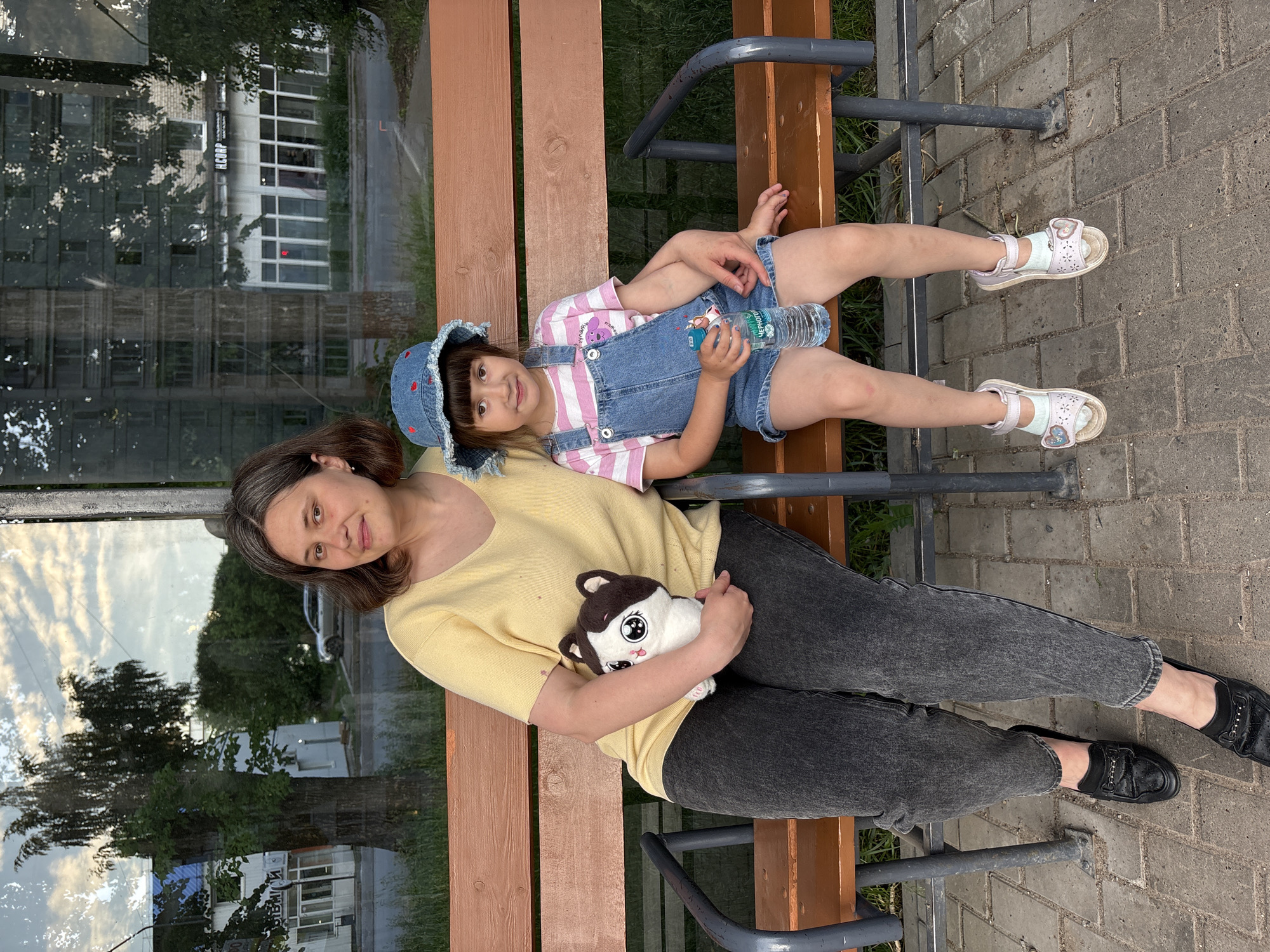 Two individuals, an adult woman and a young girl, are seated on a public bench in the city of Chaikovsky, Russia. The bench consists of brown wooden slats supported by dark gray metal frames. The ground in the foreground is paved with interlocking concrete blocks.
The adult woman, appearing to be in her 30s or 40s, has medium-length brown hair with visible gray strands. She is wearing a light yellow V-neck short-sleeved top and dark gray, faded jeans. On her feet are black loafers with a decorative gold-colored chain detail. She holds a white and brown plush toy, depicting a cat-like creature with large, expressive eyes, in her left arm.
Seated to the woman's right is a young girl, estimated to be between 4 and 6 years old. She has dark brown hair with bangs and is wearing a blue denim bucket hat adorned with red strawberry or heart-shaped embroidery. Her attire includes a pink and white striped short-sleeved t-shirt featuring a cartoon dog graphic, worn underneath blue denim overalls styled as shorts. She wears light pink sandals with a heart-shaped buckle over light blue socks. The girl holds a clear plastic water bottle, labeled "ЧЕРНОГОЛОВКА" (Chernogolovka), in her right hand. Her left arm is positioned around the woman's waist. Both individuals are looking towards the camera with slight smiles.
The background, visible through a large glass panel (suggesting a bus stop or shelter), shows an urban environment. Reflections of a partly cloudy sky are present on the glass. To the left, a large tree with light-colored bark is prominent, with a light-colored building featuring a blue roof and Cyrillic signage partially visible beyond it. A silver car is parked further down a paved road. To the right, a multi-story building with numerous windows and a commercial storefront marked with "Н.КОРП" (N.KORP) in Cyrillic can be observed. Green grass and foliage are visible in various parts of the background. The lighting suggests late afternoon or early evening.