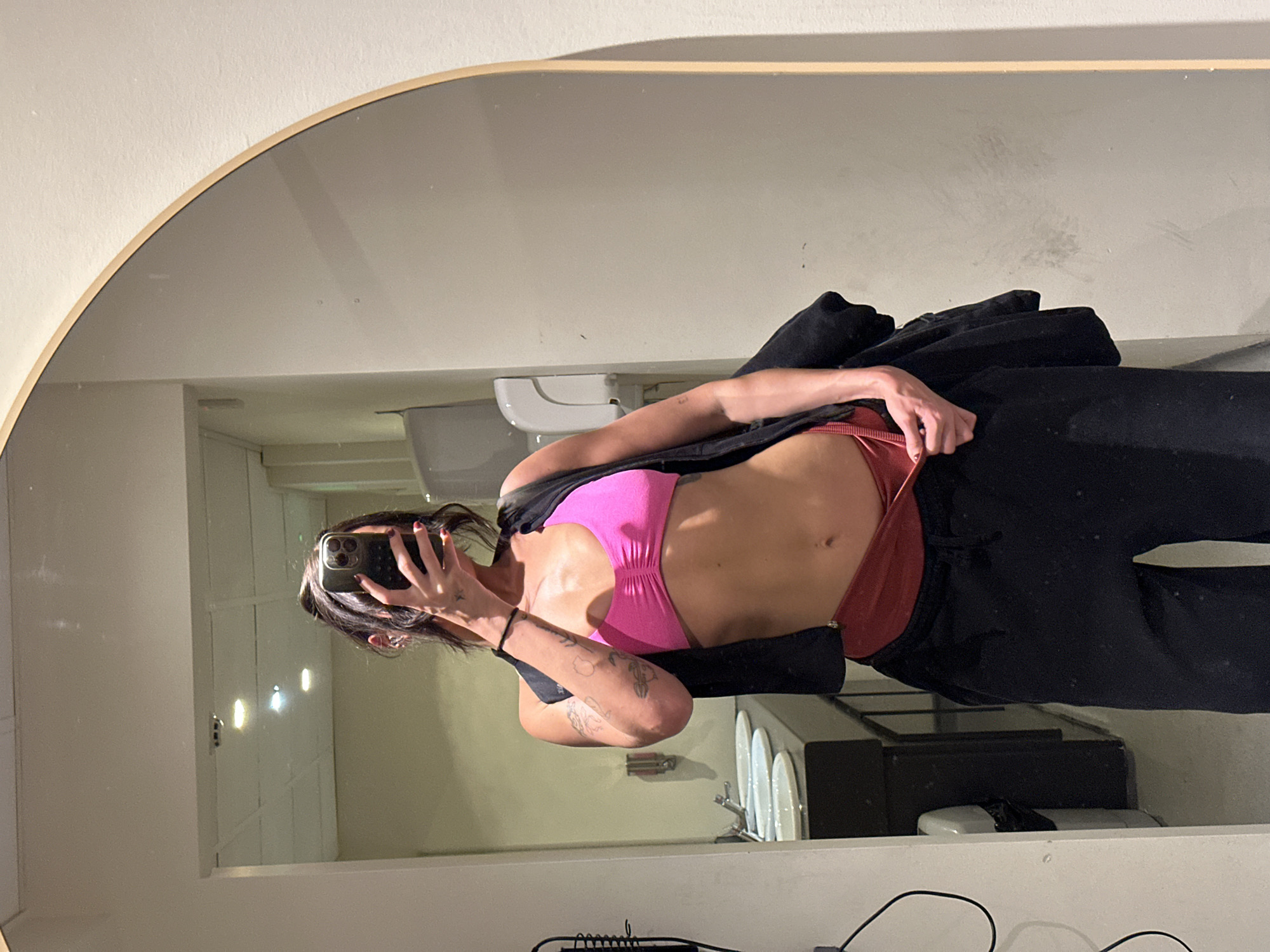 A woman is taking a selfie in a mirror, presumably in a bathroom. She is wearing a pink sports bra, red underwear, and black sweatpants with a black jacket draped over her shoulder. Her hair is dark with a streak of white. The bathroom contains a toilet, sink, vanity, and a white-tiled wall. The image appears to be rotated. There is no evidence to suggest the location is Tonbridge and Malling, United Kingdom.
