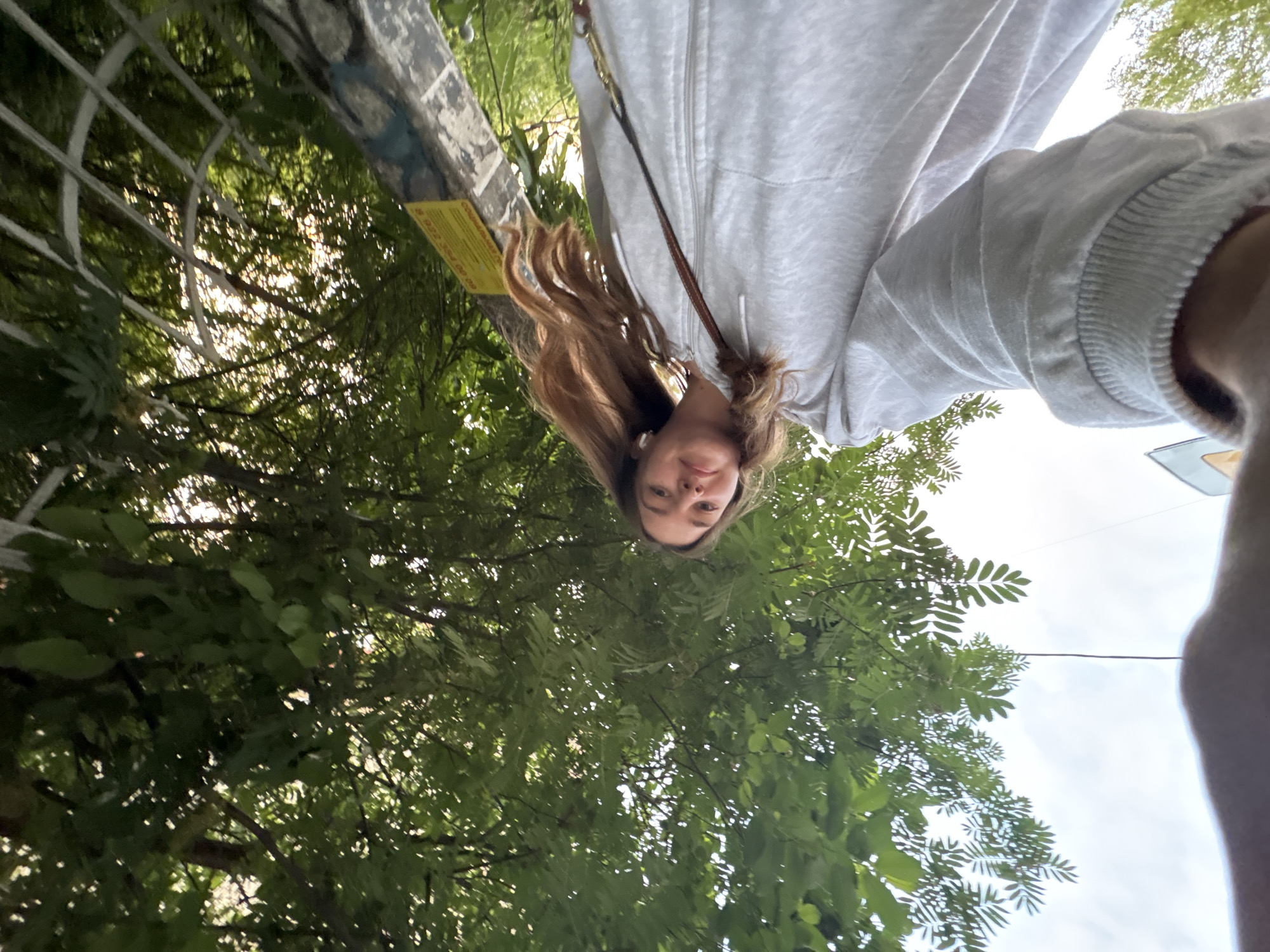 A low-angle selfie depicts a female with light brown hair and fair skin, wearing a gray hoodie. She is positioned in front of dense green foliage and trees. A white metal fence is seen in the upper left corner, next to a yellow information sign attached to the fence. The sky is visible through the leaves. The photo was taken from a position below the subject, looking upwards. No specific location within Perm, Russia, is identifiable from the image.