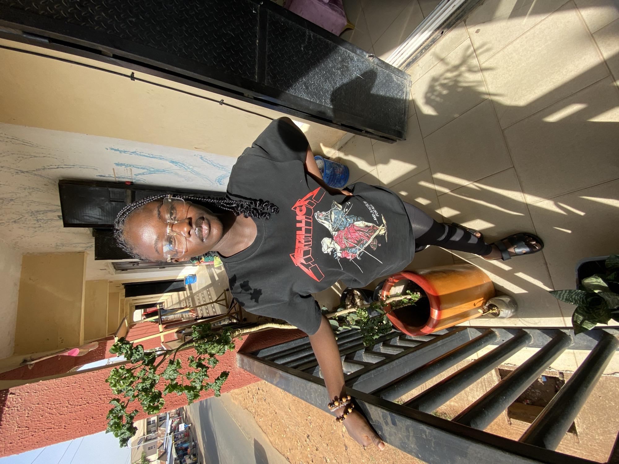 A medium shot presents a young Black woman in Bukuru, Nigeria. She wears glasses, a black t-shirt with a "Metallica" graphic, black leggings, and sandals. Her hair is styled in braids. She stands on a tiled terrace next to a black metal railing overlooking a street, with a view of buildings and traffic below. A clay pot sits beside her, and plants are visible in the background. The scene is lit by bright sunlight, casting shadows across the terrace.