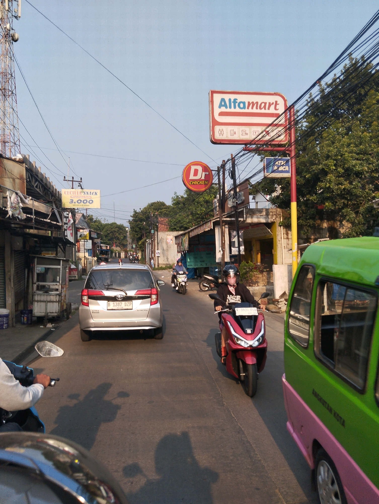 The image captures a street scene in Sukaraja, Indonesia, with a focus on traffic and commercial signage. A silver Toyota Avanza, license plate B 1387 DZP, is prominently featured in the foreground, driving on the right side of the road. To the right of the Avanza, a green and pink vehicle, possibly a small van or bus, is also in motion.
In the middle ground, a person on a red scooter, wearing a black jacket and a white face mask, is visible. Further down the street, another scooter rider can be seen. The environment is lined with buildings housing various businesses. A prominent sign for "Alfamart" is visible, along with a red circular sign for "Dr CHICKEN." A yellow sign indicates "CECILIA SNACK" with a price of "3.000". There are also signs for "ATM BCA."
The street is paved with asphalt, and there are overhead power lines crisscrossing the sky. The lighting suggests it is daytime, with the sun casting shadows on the road. The overall impression is a bustling urban street with typical Indonesian traffic and commercial activity.