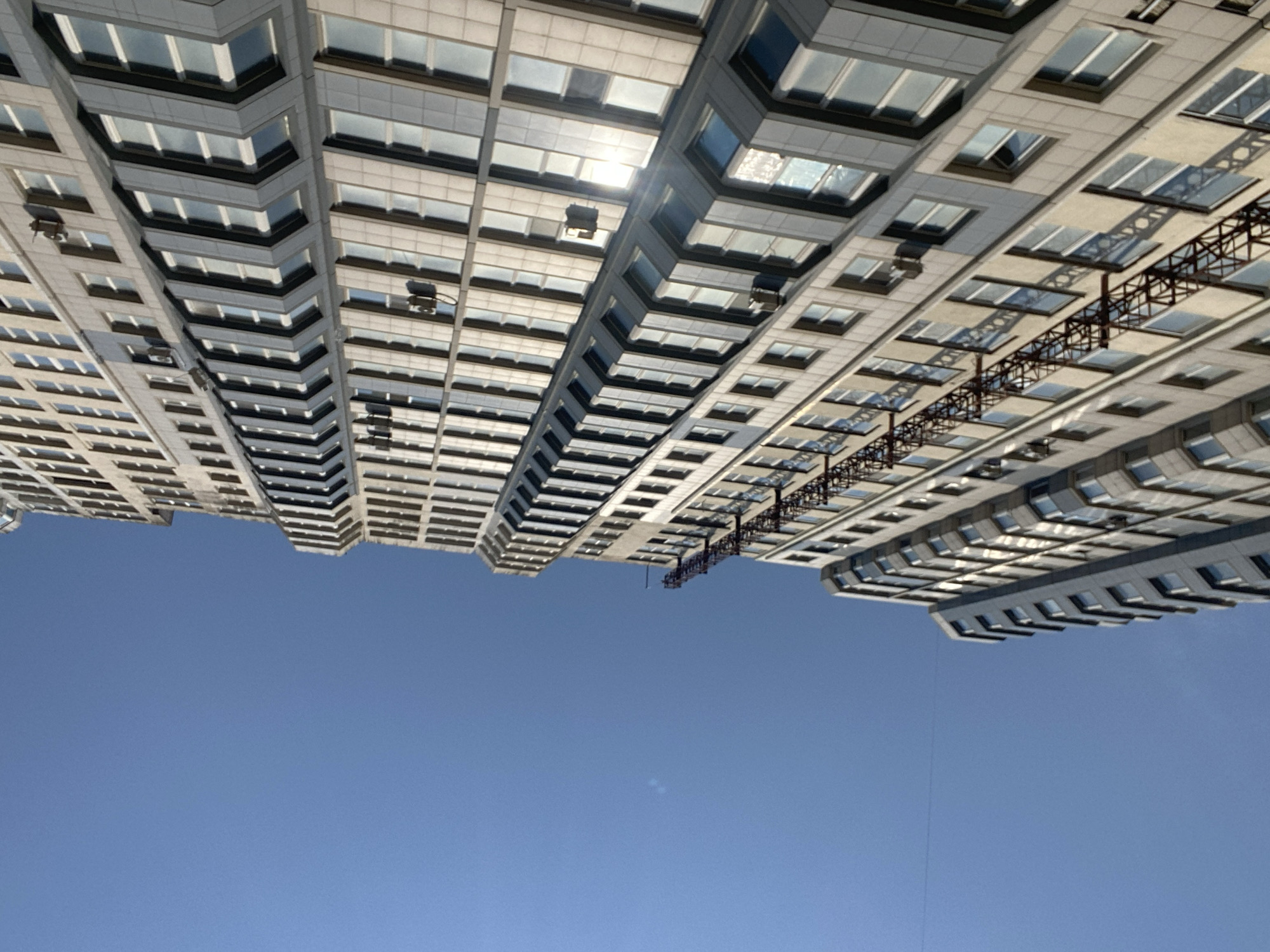 The image, taken from a low angle looking upwards, displays the facade of a modern, multi-story residential building against a clear blue sky. The building, oriented vertically in the frame and slightly tilted, exhibits a repetitive pattern of windows with light-colored frames and reflective glass, suggesting a contemporary architectural style. The exterior material appears to be a combination of light-colored panels and darker gray accents around the windows. Several air conditioning units are visible mounted on the exterior walls. A prominent feature of the building is a dark, lattice-like structure, possibly a fire escape or external scaffolding, running vertically along a section of the facade. The overall scene conveys an urban environment, likely a city like Chornomorsk, Ukraine, with a focus on the building's structure and its interaction with natural light. There are no people or any discernible actions taking place within the frame.