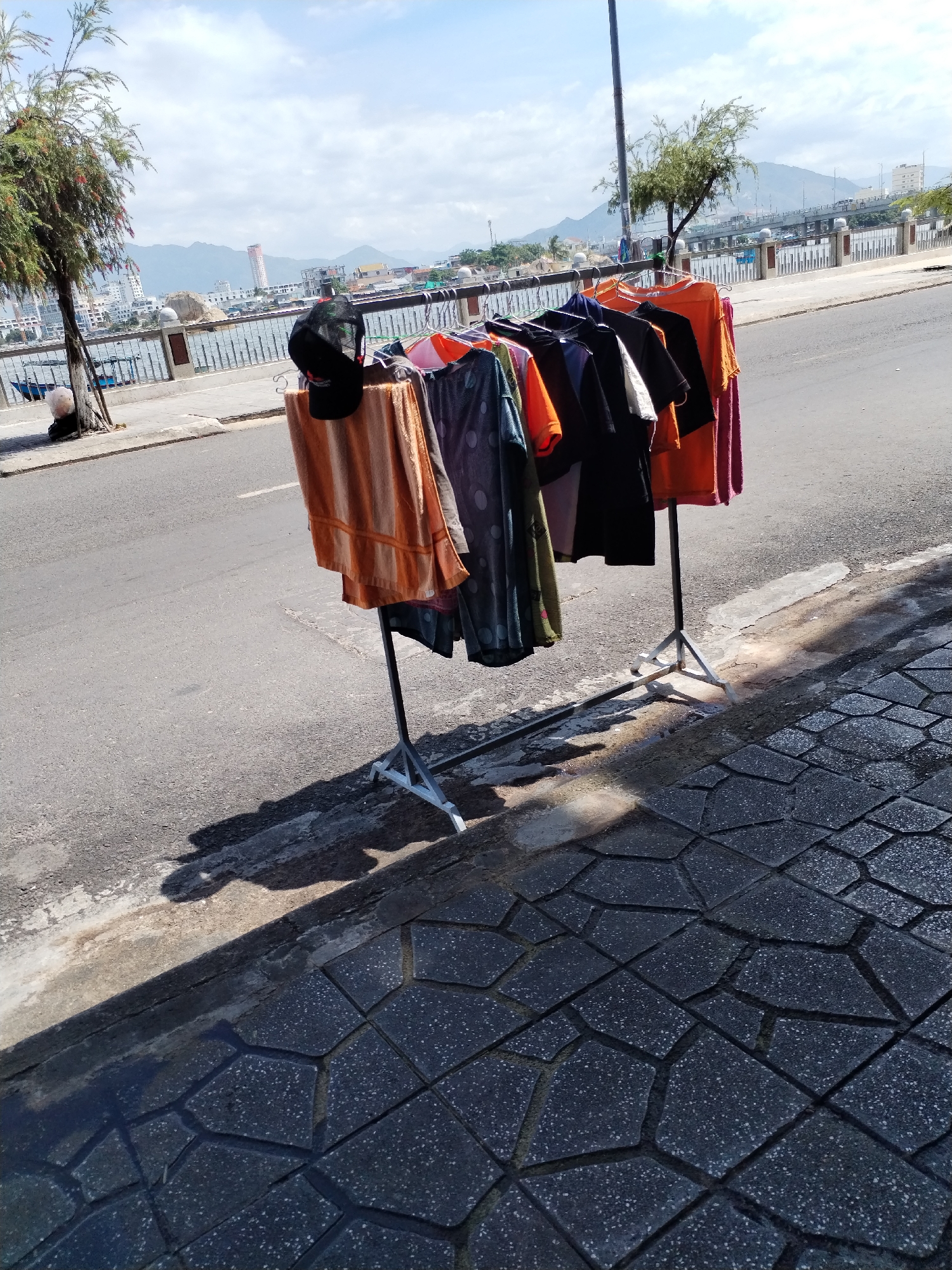 In the city of Nha Trang, Vietnam, along a paved walkway adjacent to a road, a clothes rack displays an assortment of folded clothing items and t-shirts. The clothing includes items in orange, purple, black, and green, with some t-shirts featuring polka dot patterns. A black baseball cap rests on top of the rack. The background reveals a city skyline with buildings and mountains, a body of water with a small boat, and a bridge. The scene is illuminated by sunlight, casting shadows from the clothes rack and a nearby tree onto the patterned pavement.
