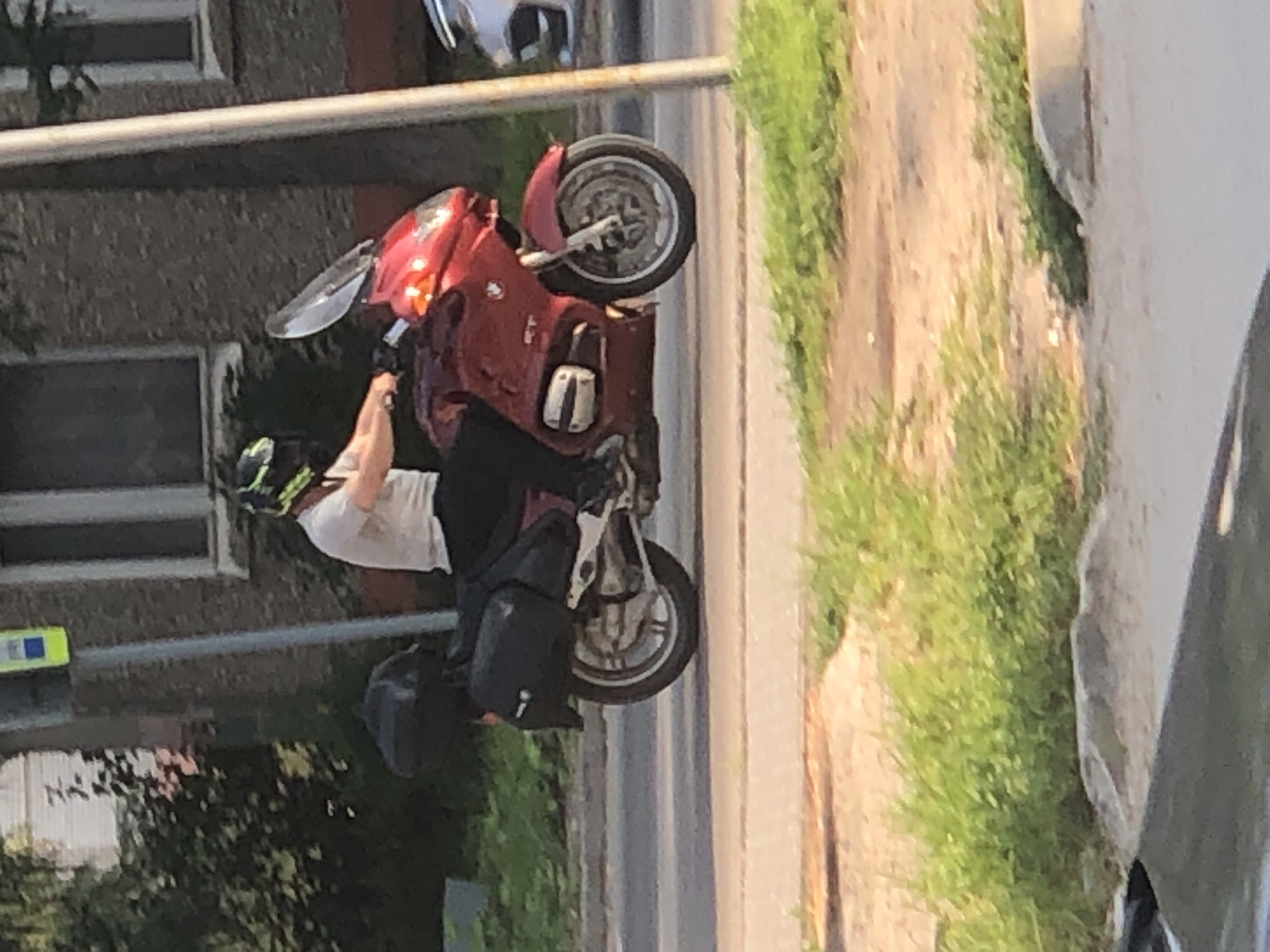A person wearing a white t-shirt and dark pants is riding a red motorcycle with two black saddlebags. The motorcycle has a large windshield. The person is wearing a helmet with green and black accents. The motorcycle is on a paved road with a grassy area to the left and a building with windows to the right. A gray pole with a yellow and blue sign is visible behind the motorcycle. The scene appears to be an outdoor urban environment in Solikamsk, Russia.