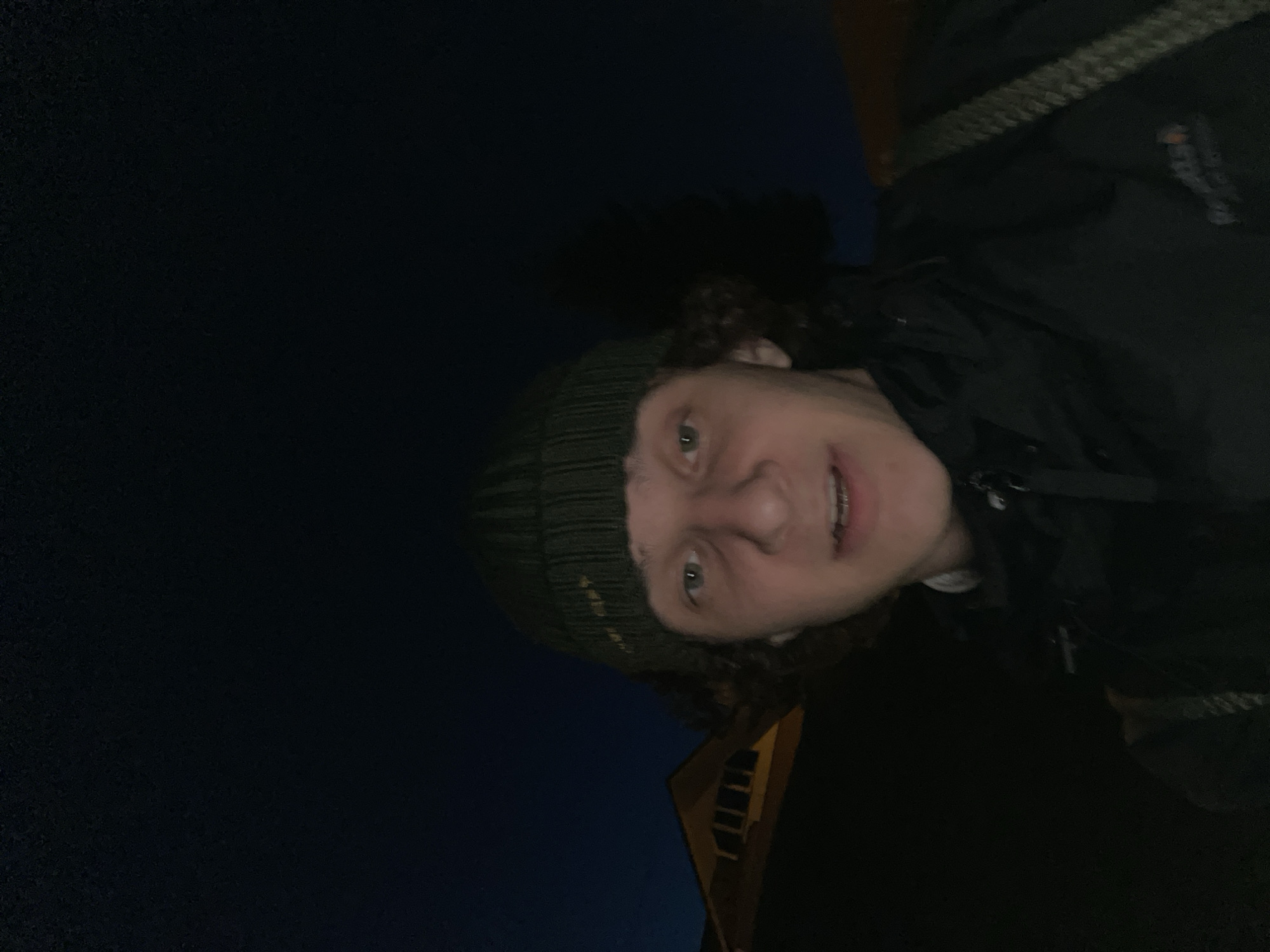 A person wearing a dark green beanie and a dark jacket is posing for a selfie. The background is dark, suggesting it is nighttime. In the lower center, a section of a building with lit windows is visible. The location is potentially Krasnoyarsk, Russia.