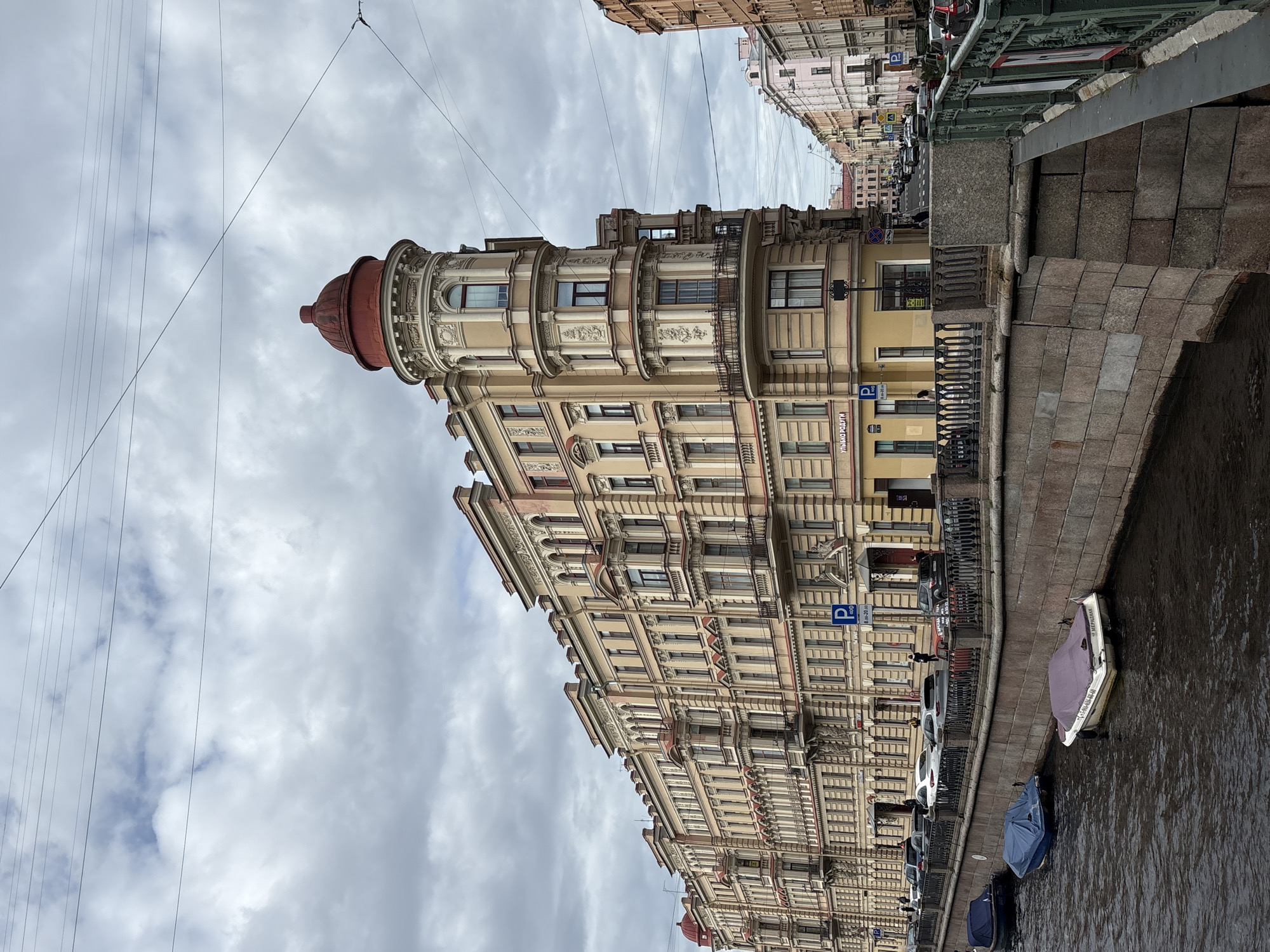 This is a street view photograph of a canal in Saint Petersburg, Russia. The canal is lined with a stone embankment and an ornate metal railing. Several boats are moored along the embankment, some covered with tarps. Across the canal, a large, ornate yellow building with a red domed tower dominates the scene. The building features multiple balconies, intricate architectural details, and numerous windows. Further down the street, other buildings with varying architectural styles are visible. The sky is overcast with clouds. Cars are parked along the street and there are a few pedestrians visible. A street sign with Russian text and a blue parking symbol is present on the building. The photograph is taken from an eye-level perspective.