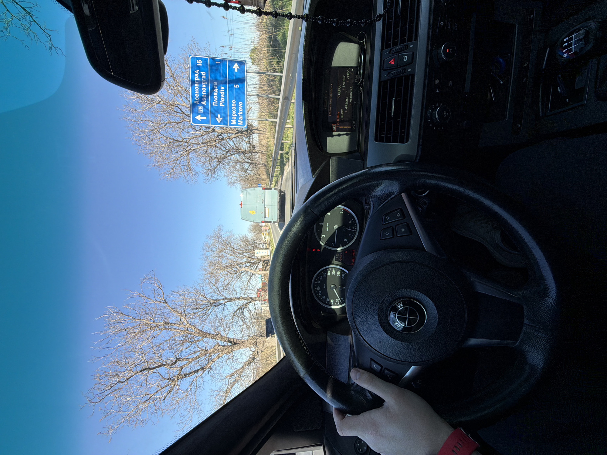The photograph shows a person driving a car on a road near Plovdiv, Bulgaria. A road sign indicates directions and distances: Asenovgrad is 16 kilometers in one direction, Plovdiv is 1 kilometer in another, and Markovo is 5 kilometers in yet another direction. The car's dashboard is visible, showing a radio station display and the steering wheel with a "BMW" badge. A hand, wearing a red wristband, is visible on the steering wheel. The exterior environment consists of trees without leaves under a blue sky, suggesting a colder season. A white vehicle is also visible ahead on the road.