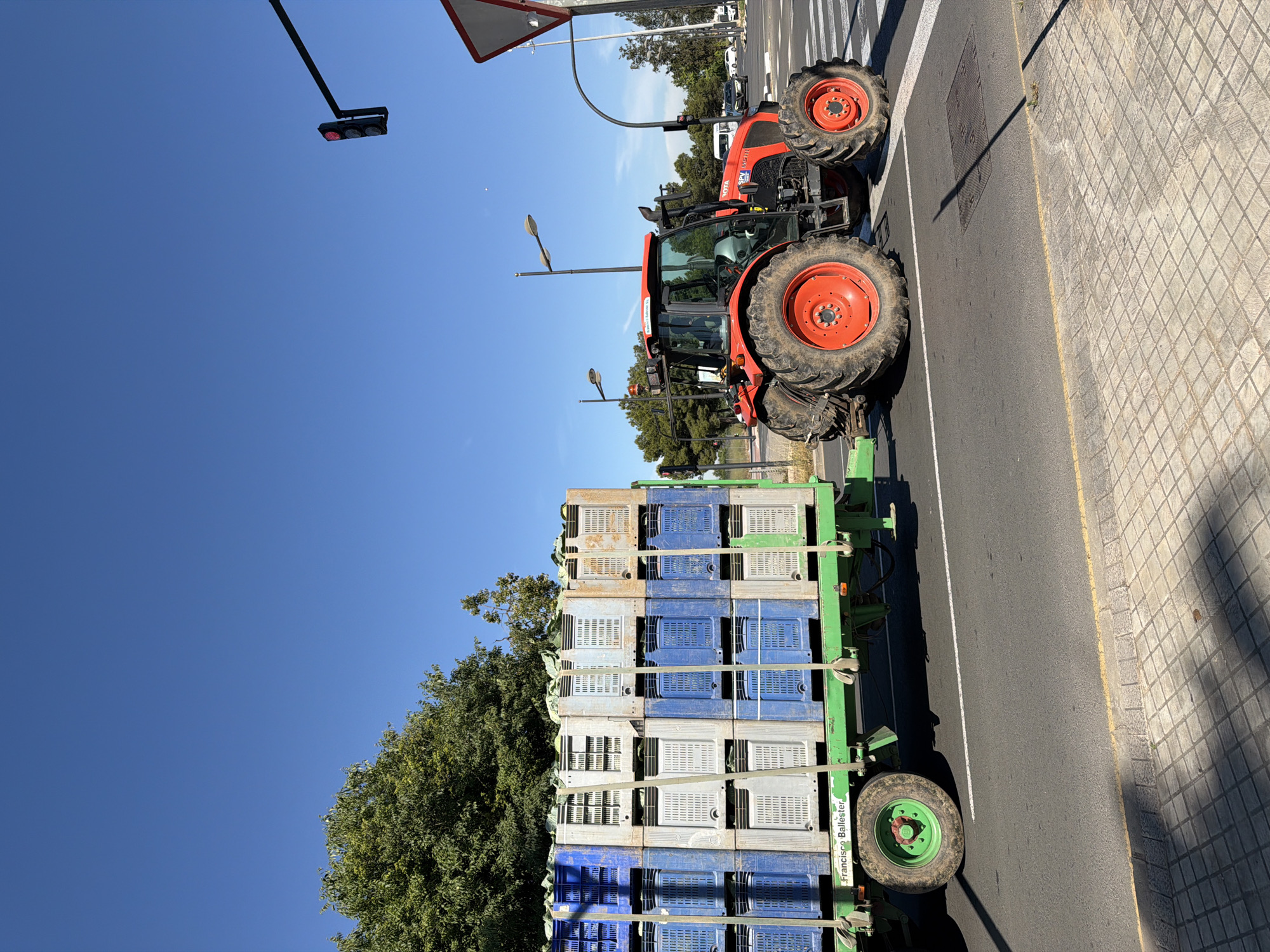 A medium, eye-level shot depicts an orange tractor hauling a green trailer filled with stacked blue and white plastic crates, parked on a paved street. The tractor is on the right side of the road, near a crosswalk and a street sign. The crates appear to be empty. Behind the trailer are trees and more streetlights. The sky is clear and blue, with a red traffic light visible in the background. The text "Francisco Ballester" is printed on the trailer. The location is in Valencia, Spain.