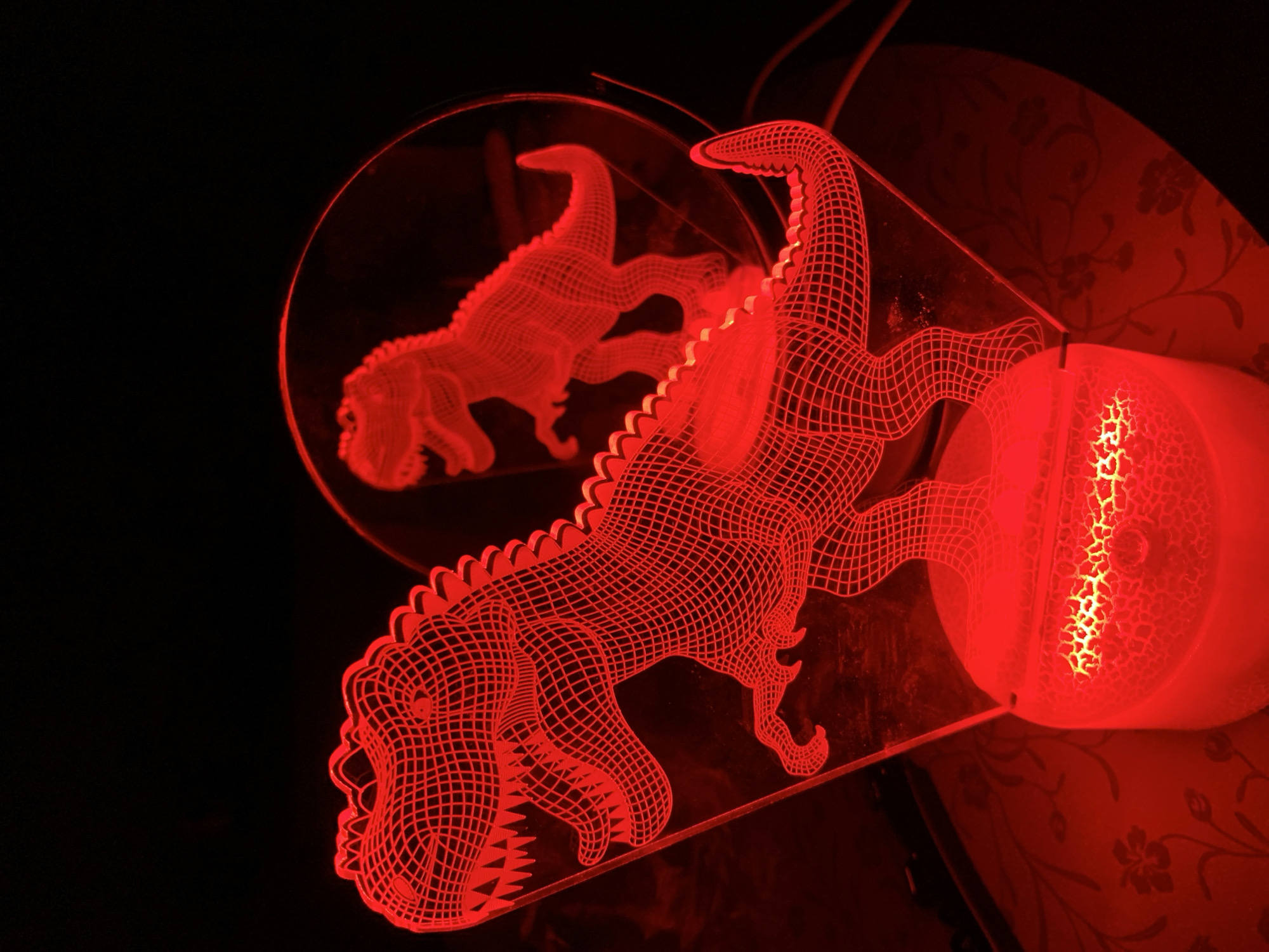 The image shows a close-up of a red 3D illusion lamp shaped like a Tyrannosaurus Rex. The lamp is lit, casting a vibrant red glow and a reflection of another T-Rex on a circular mirrored surface behind it. The lamp sits on a textured surface, possibly a table, with a floral pattern visible in the red light. The background is dark and out of focus. There are no people or other animals in the image. The location in the city of Chaikovsky, Russia, is not determinable from the image.