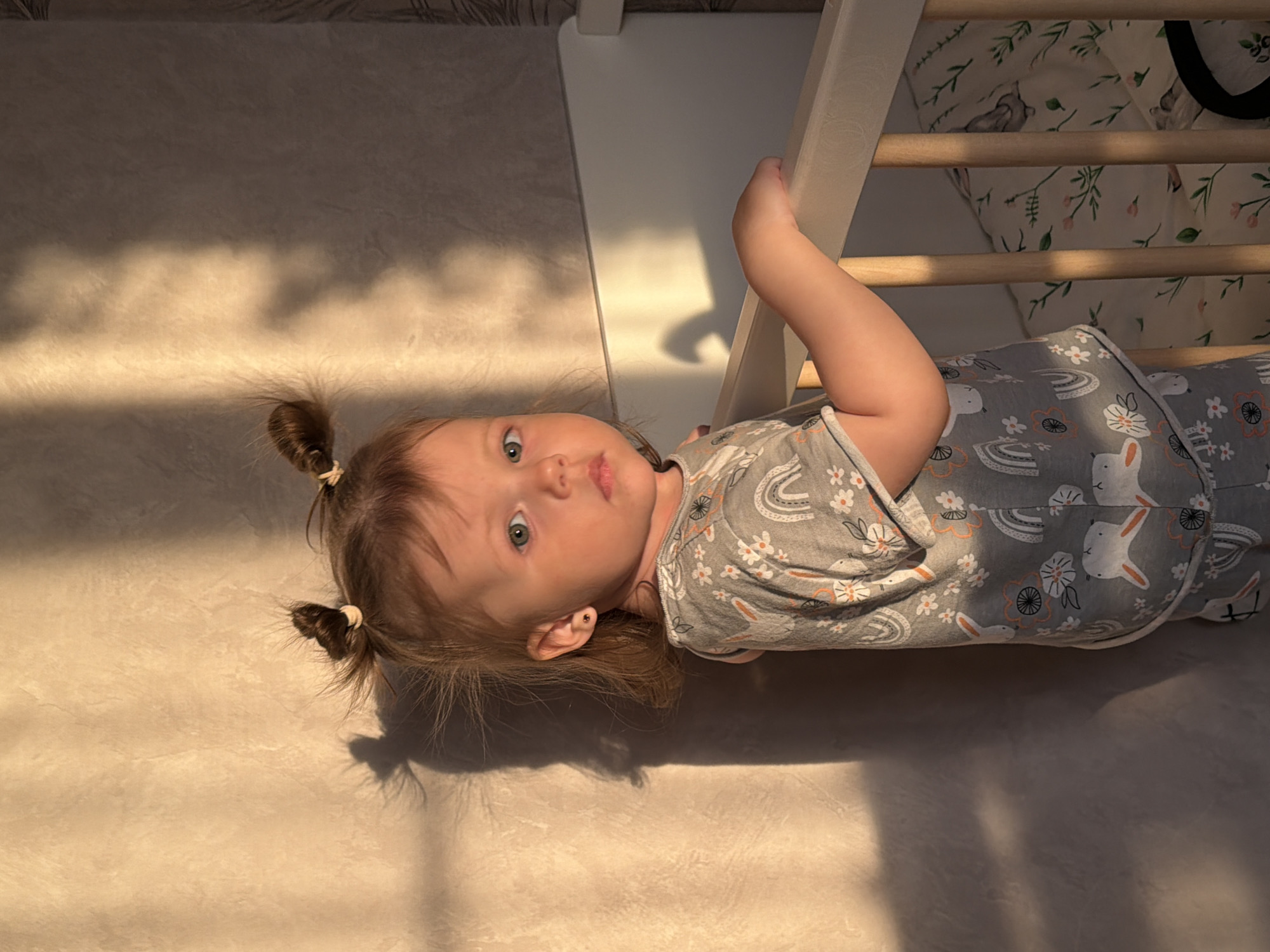 This image captures a young child lying on their back in an indoor setting, looking directly at the camera with a neutral, curious expression. The child has light brown hair styled in two small pigtails secured with light elastic bands and strikingly blue-green eyes. Small stud earrings are visible in their ears. They are wearing a grey garment, likely a romper or pajamas, patterned with white rabbits, rainbows, and small flowers in white and orange.
The child's right arm is raised, holding onto a white wooden slat of what appears to be a Pikler triangle or a similar indoor climbing frame/crib. The frame features several horizontal wooden rungs. Behind the frame, in the upper right quadrant of the image, visible bedding or cushions are patterned with green leaves, pink flowers, and rabbits, similar to the child's clothing. The background in the upper left shows a patterned wall.
The setting is a room with a light-colored floor, possibly carpet or textured tile. The space is illuminated by prominent golden shafts of sunlight streaming in, casting distinct, elongated shadows across the floor and furniture. This warm, directional lighting suggests either early morning or late afternoon.
The overall impression is one of a peaceful domestic scene in Reshetnikovo, Russia. There is no visible text within the image.