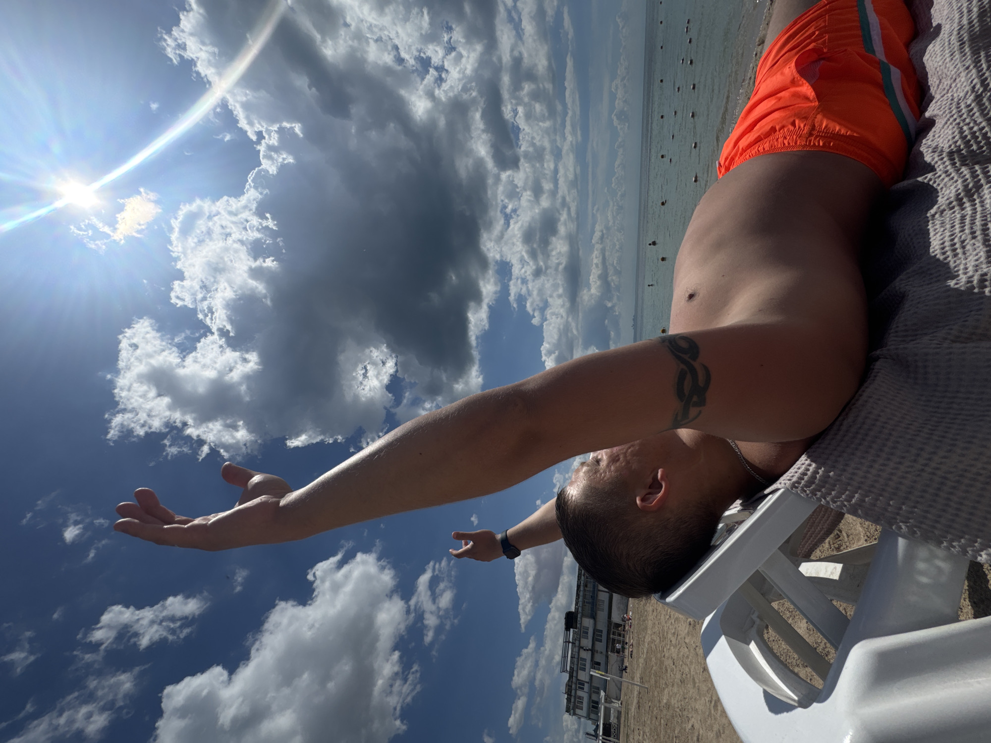 A low-angle view shows a man lying on a white plastic beach chair at a beach in Yevpatoriya, Ukraine. He is wearing orange swim trunks with white and green stripes, and he is lying on a gray towel. He is shirtless and has his arms outstretched, with one hand raised toward the bright sun. The sky is blue with fluffy white and dark clouds. The water is calm and there are people swimming and standing in the distance. In the background, there is a building on the beach and the sand stretches out to the left. The scene is sunny and the man appears to be relaxing.