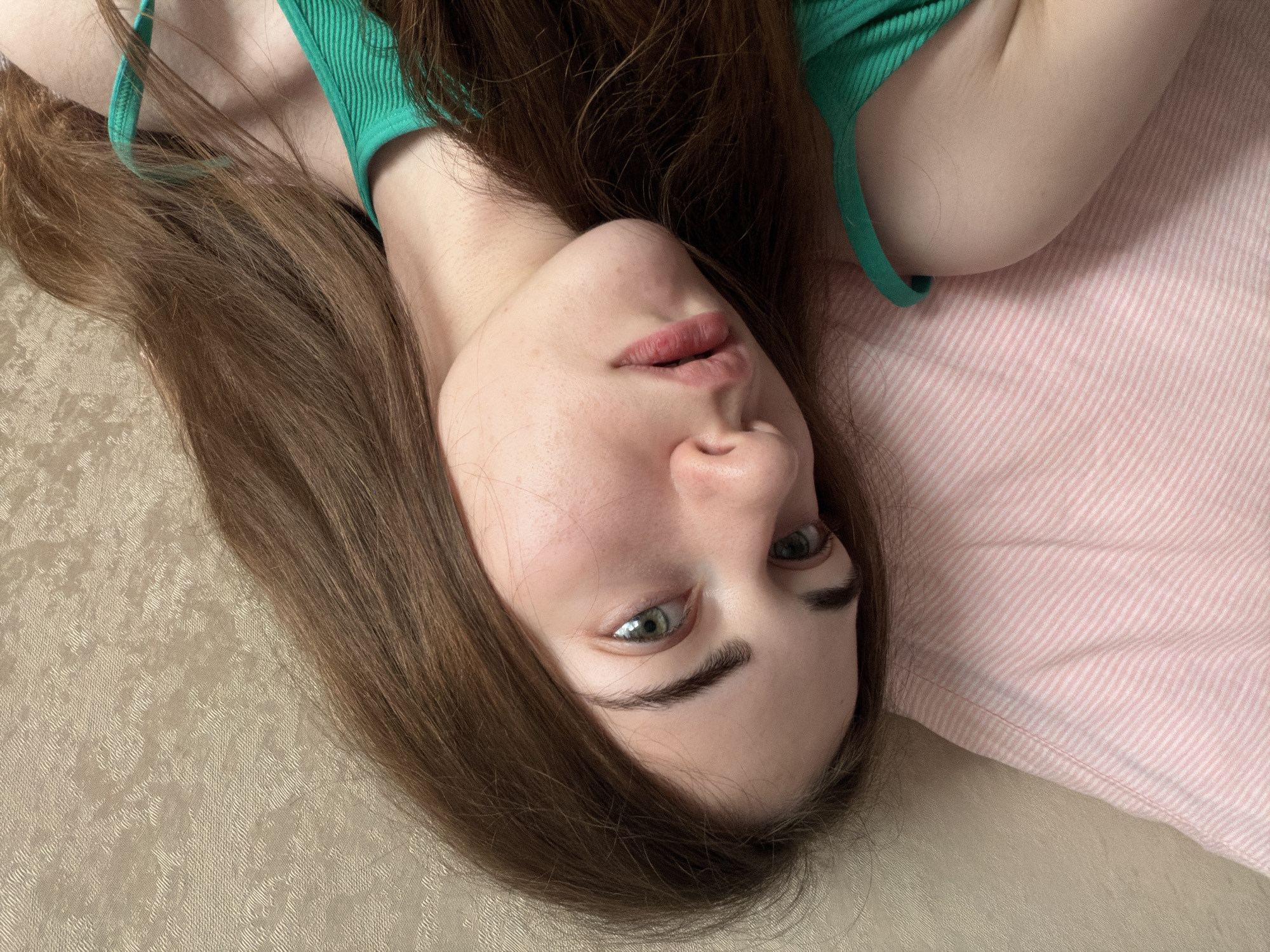 The image shows a close-up, overhead shot of a young woman with long, brown hair lying on a pink and white striped pillow. She is wearing a green tank top. Her head is tilted slightly, and she is looking towards the camera with a neutral expression. The background is a textured, light-colored surface, possibly a wall or headboard. The image appears to be taken indoors. There is no information in the image to indicate a specific location in the city of Tyumen, Russia.