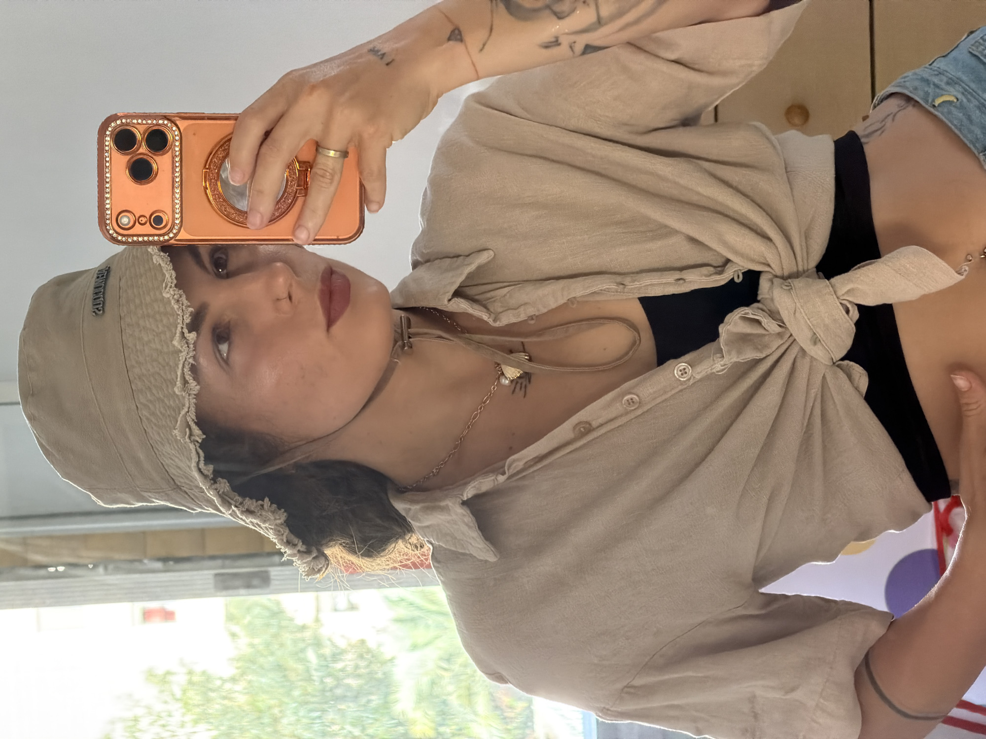 A person is depicted taking a mirror selfie, showing their upper body and face. The original image is rotated 90 degrees counter-clockwise.
**Subjects:** The primary subject is an individual, likely female, with medium-toned skin and dark hair, visible from the chest up. She has a neutral expression and is wearing dark pink lipstick.
**Objects:** She is holding an iPhone in her right hand, encased in a decorative, peach/rose gold colored case featuring rhinestones around the camera lenses and a circular ring holder on the back. She wears a gold-colored ring on her right ring finger. Her attire includes a light beige or tan bucket hat with frayed edges, featuring mirrored text (possibly a brand name, like "JENTINLIV" when read mirrored) on the front. She is wearing a light beige, linen-like button-down shirt, tied at the front to expose her midriff, with a black top or bra visible underneath. Multiple necklaces adorn her neck, including a thin gold chain with small charms and a thicker, light-colored choker-style necklace with a metallic detail. Small stud piercings are visible on her right ear. Parts of blue denim clothing (likely shorts or jeans) are visible on her right side. Several small tattoos are visible on her left arm/shoulder, including some text or symbols, and a band-like tattoo on her right upper arm.
**Scene and Setting:** The photo is taken indoors, reflected in a mirror. Behind the person, a large window is visible, letting in bright natural light. Through the window, green foliage (trees or bushes) and a bright sky are seen, suggesting daytime and sunny weather outside. To the right of the reflection, part of an indoor space is visible, including what appears to be wooden furniture or a wall, and colorful, abstract shapes (red, purple, yellow circles) in the lower right background.
**Activity:** The person is engaged in the activity of taking a mirror selfie.
**Notable Details:** The detailed and embellished phone case is a prominent feature. The visible tattoos and various accessories add to the person's style. The natural light indicates it is daytime.
