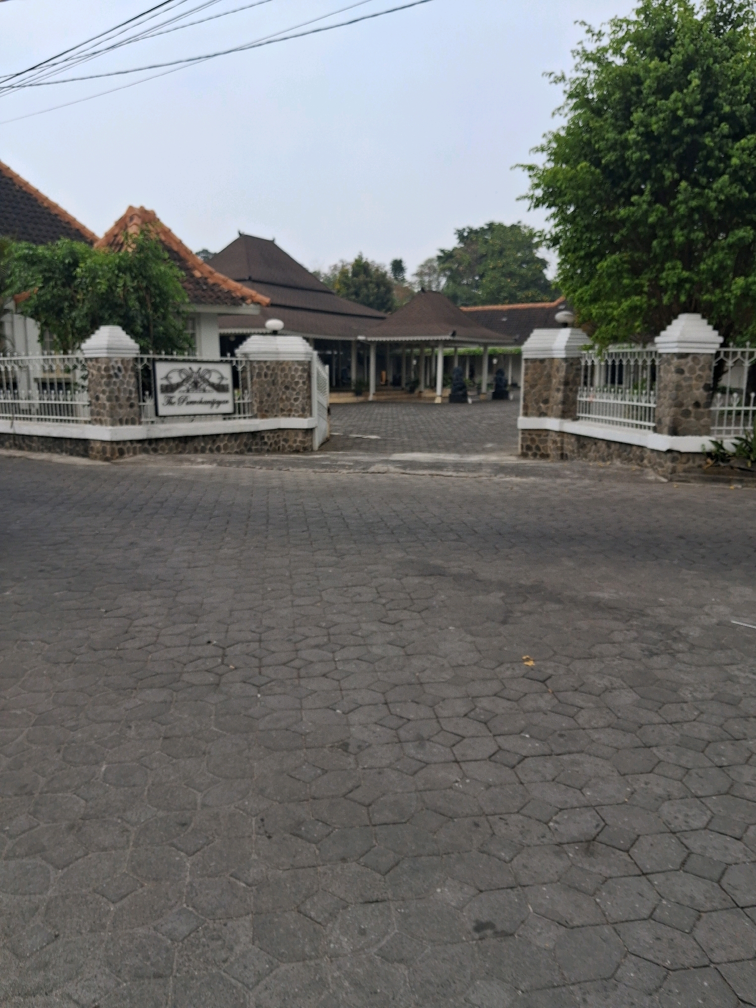 A paved street leads to a white fence and gate with stone pillars. Behind the fence is a large building with a dark, multi-tiered roof and white pillars supporting an open-air walkway. The building is surrounded by trees and lush greenery. A sign is visible on the fence, reading "The Pamantjingan". The sky is overcast and the overall scene suggests a well-maintained property in a tropical or subtropical climate. This is likely the entrance to The Pamantjingan, a heritage building located in Surakarta, Indonesia.
