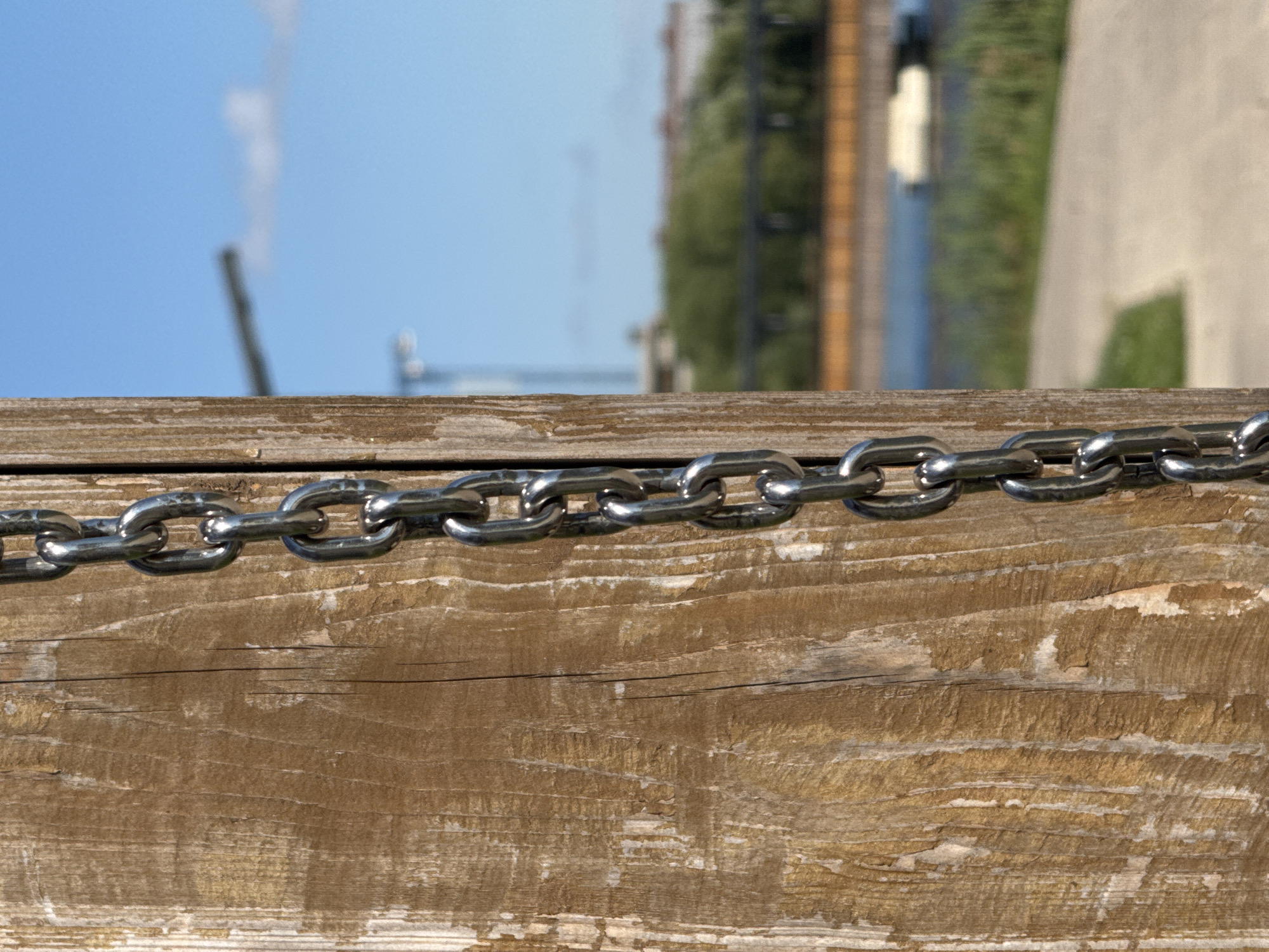 A close-up, vertical shot showcases a metal chain suspended alongside a weathered wooden post in Kommunarka, Russia. The chain, made of interlocking metallic links, hangs prominently in the foreground, casting subtle shadows on the rough, textured surface of the post. The background is softly blurred, revealing a tranquil scene with a body of water, lush green vegetation, and a distant bridge. The sky above is a clear, vibrant blue, with a faint white cloud adding a touch of contrast. The composition is dominated by the stark juxtaposition of the man-made chain and post against the natural backdrop, suggesting an outdoor recreational or public space. There are no people or animals visible in the image.