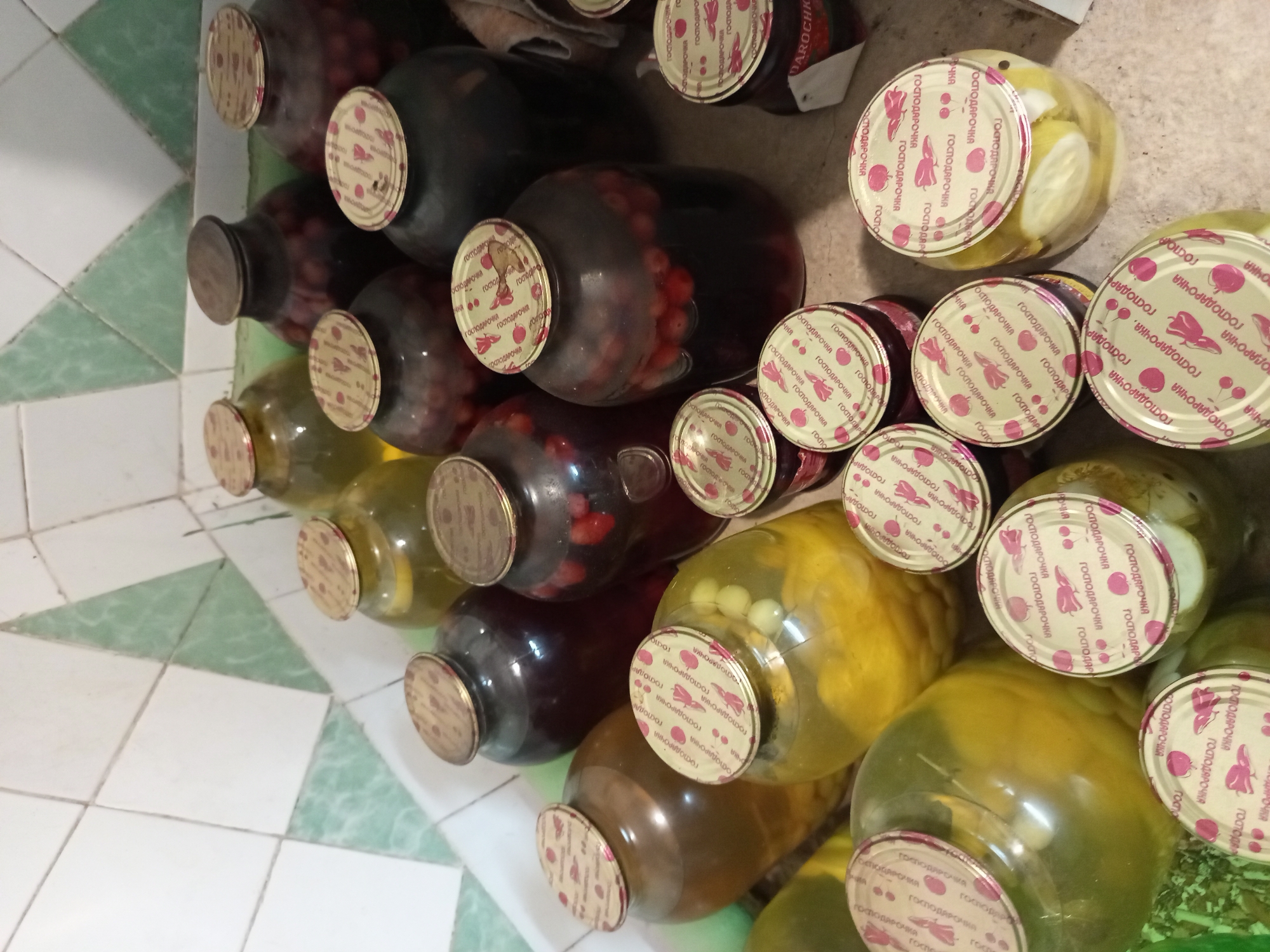 A collection of glass jars filled with preserved food items sits on a tiled surface. The jars vary in size and contain different contents, including what appears to be dark berries, yellow fruits or vegetables, and possibly pickled cucumbers. Most of the jars are sealed with patterned metal lids, many of which have Cyrillic writing. The background is a tiled wall with a repeating pattern of white and green geometric shapes. The lighting is somewhat dim, suggesting an indoor, possibly storage, environment. The scene is devoid of people or any discernible activity. The context of Tiraspol, Moldova, is not directly evident from the visual information in the image itself; it is an external piece of information.