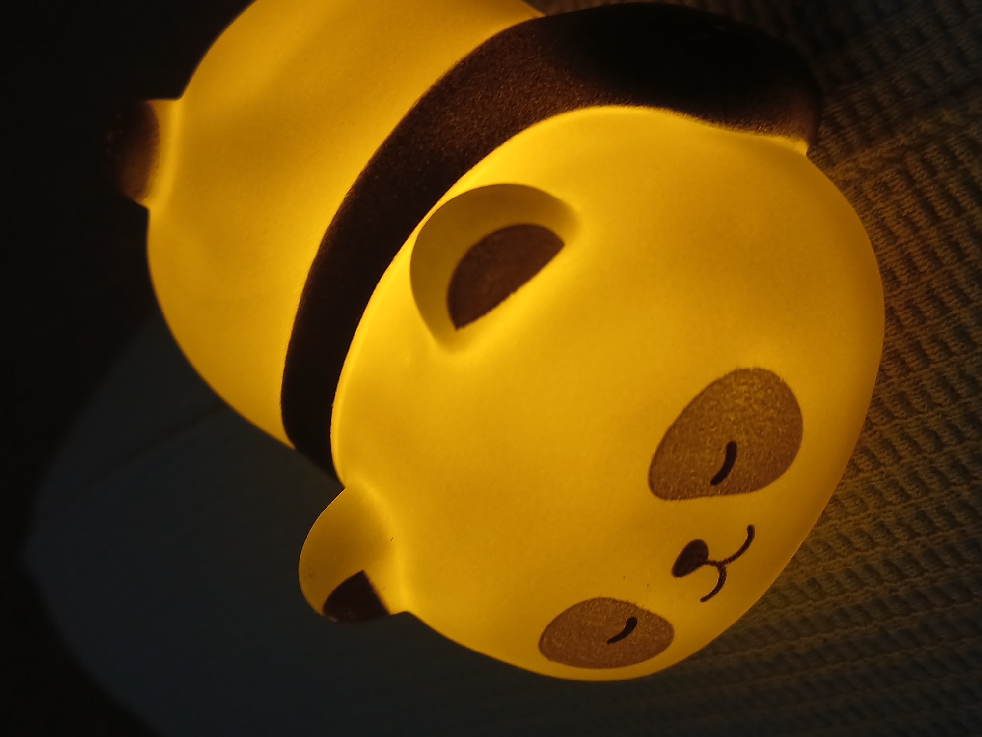 The image displays a glowing, panda-shaped lamp or nightlight, lying diagonally across the frame. The object emits a soft, warm yellow light, making it the central illuminated feature in a dimly lit indoor environment. The panda's body is a smooth, luminous yellow, contrasted by dark brown or black details that define its features: rounded ears, eye patches, and a prominent stripe wrapping around its torso. Its face shows simple, stylized closed eyes and a small nose and mouth, conveying a peaceful or sleeping expression.
The illuminated panda rests on a dark, textured surface, possibly a woven fabric, blanket, or carpet, which is visible to the right and partially at the top of the image. This surface has a patterned, slightly ribbed texture that softly catches the glow from the lamp. The background to the lower left is dark and indistinct.
No activity or event is taking place, as the image is a static portrait of the object. The noticeable light emanating from the panda suggests it is nighttime or that the room is otherwise dark, highlighting its function as a source of comfort or ambiance. There are no people, visible text, or specific outdoor weather conditions depicted. While the location context is Bengaluru, India, there are no explicit visual cues within the image itself to confirm this. The overall impression is one of calm and warmth.