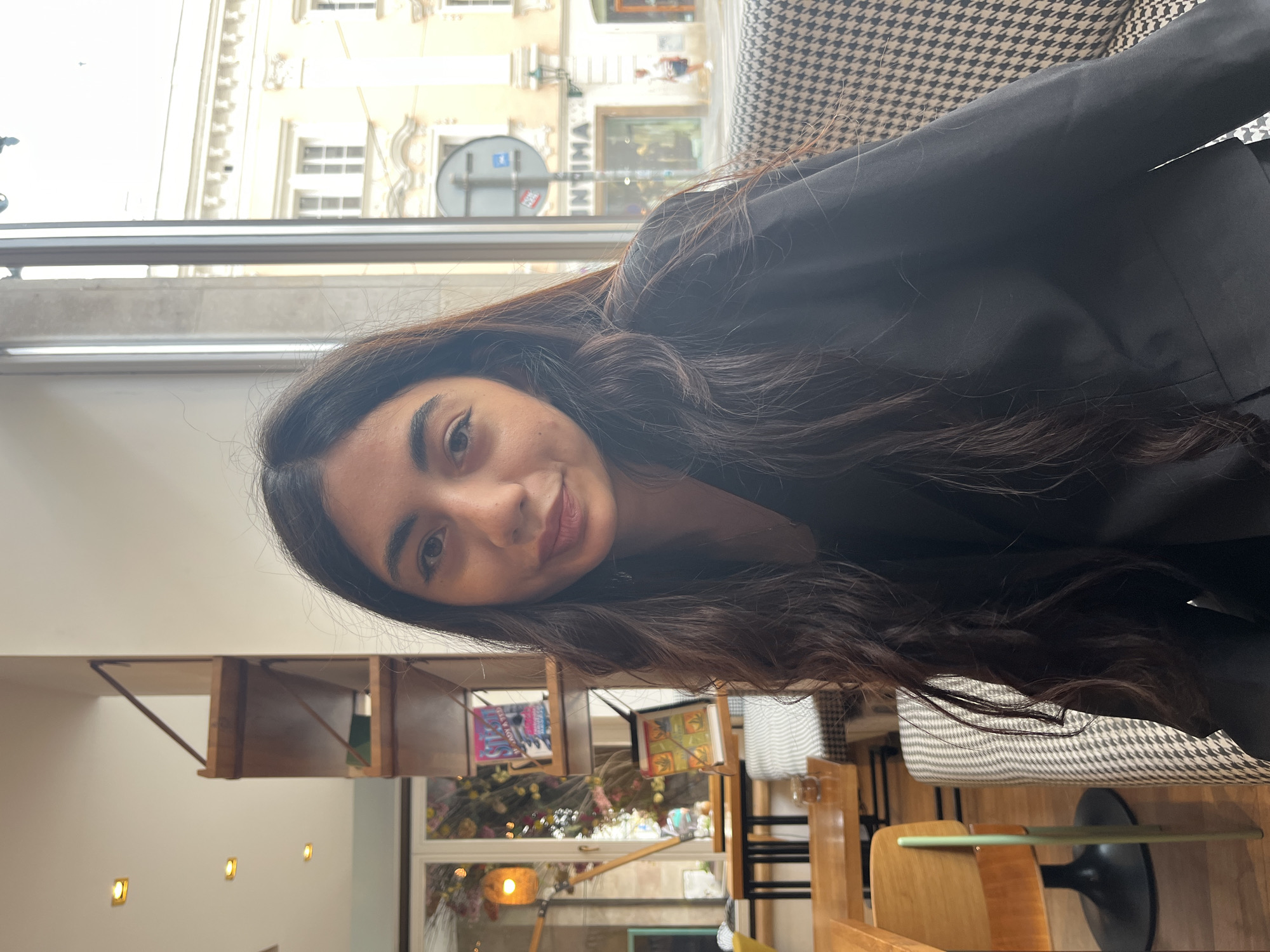 A young woman with long, dark, wavy hair is seated in a cafe in Pilsen, Czechia. She is wearing a black blazer. The background shows a window with a view of a European city street, with buildings and a sign that reads "INTIMA". There are shelves with books and decorative items behind her. The cafe has wooden tables and chairs. The woman is looking directly at the camera.