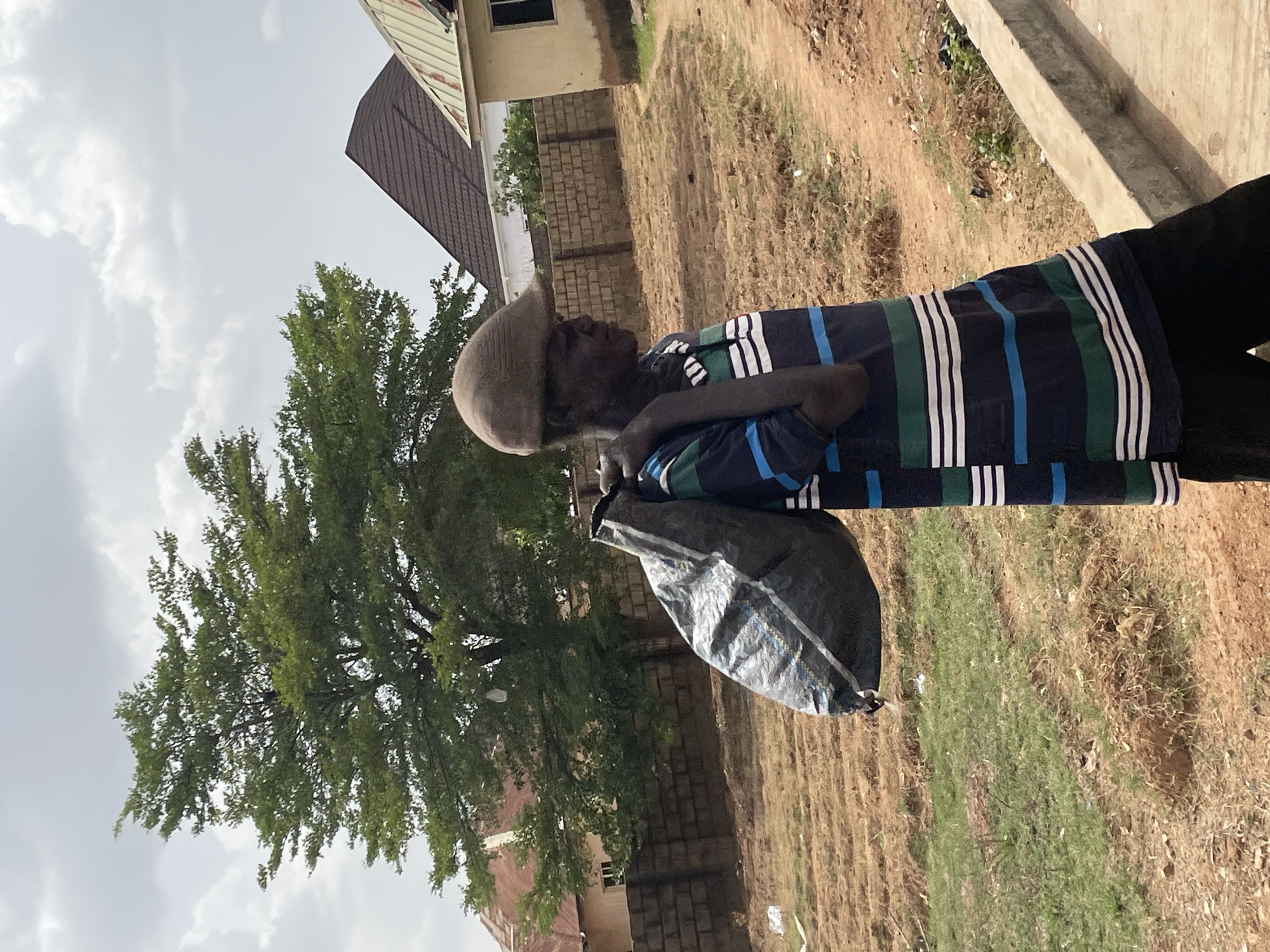 An elderly man in Bar Jirgi Summa, Nigeria, wearing a striped shirt and a hat, carries a plastic bag over his shoulder. The environment consists of a partially vegetated, grassy area with buildings and trees in the background. The sky is overcast.