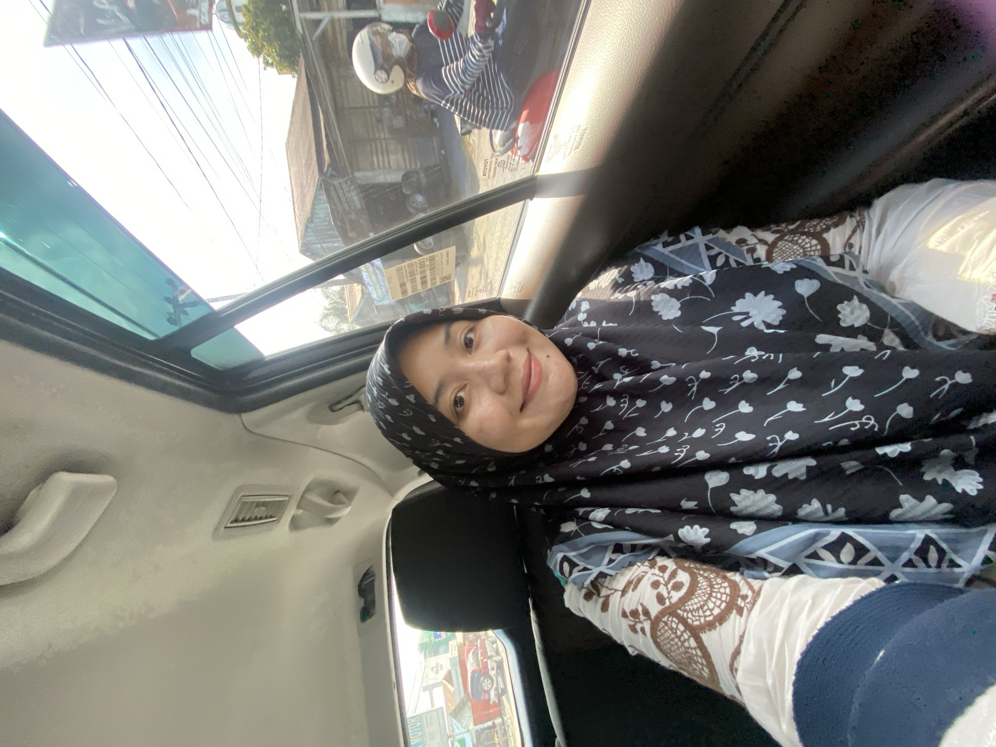 A woman wearing a hijab and patterned clothing is sitting in the backseat of a vehicle. She is looking towards the camera and smiling. The window of the car shows a street scene with buildings, a motorcycle with a rider, and power lines. The overall impression is a candid snapshot taken during a car ride in an urban setting. The scene is consistent with an outdoor environment in the city of Banjarbaru, Indonesia, given the architectural style of the buildings and the presence of typical Indonesian street elements.