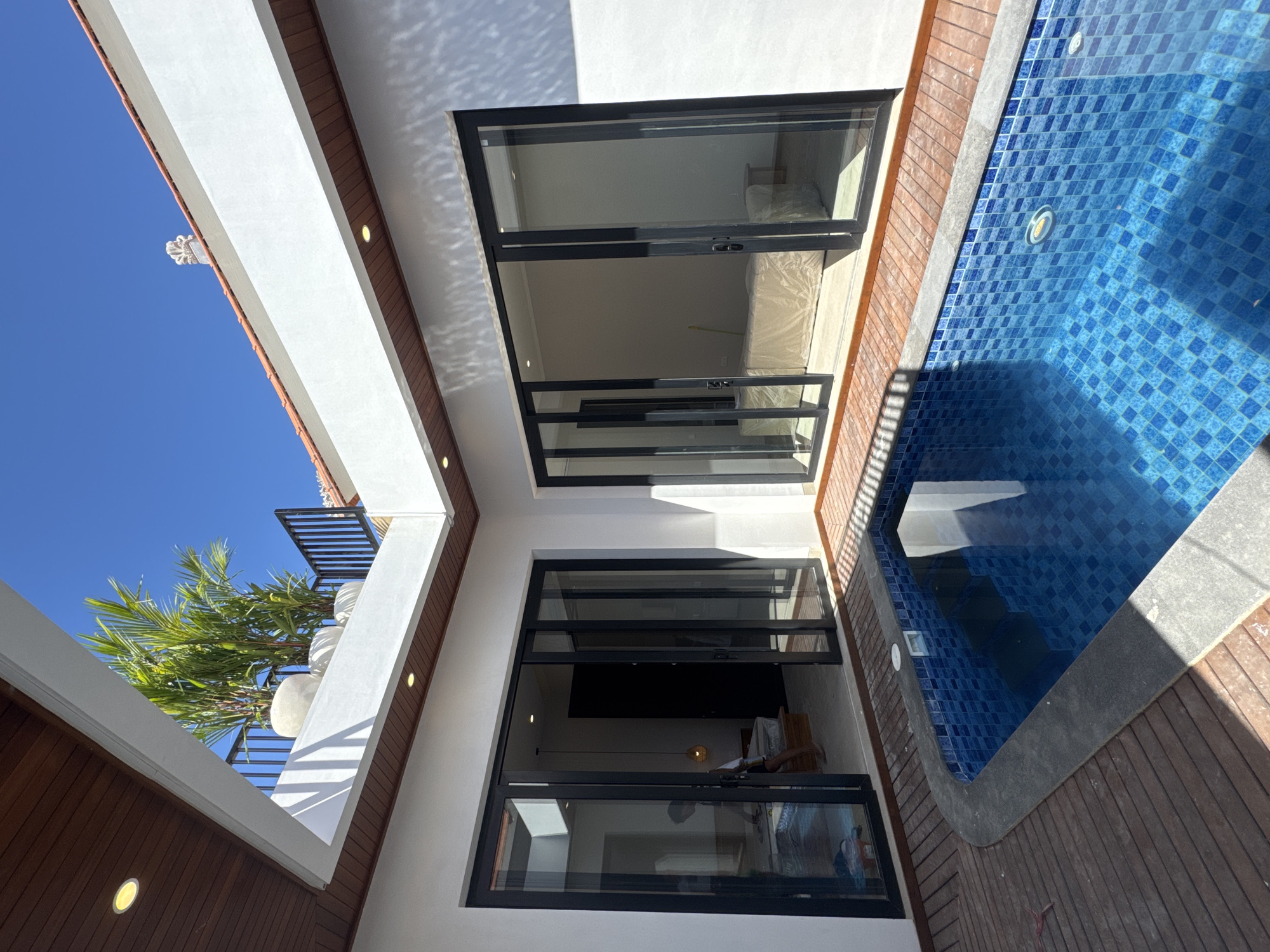 The image captures an upward-angled view of a building facade in Denpasar, Indonesia, juxtaposed with a section of a pool. The building features white walls, dark brown wooden accents, and large, black-framed glass doors that open into interior spaces. One room reveals furniture covered in protective material, suggesting recent construction or renovation. The pool is lined with blue mosaic tiles and is bordered by a wooden deck. A blue sky and palm trees are visible in the background, adding to the tropical setting. No people are visible in the scene.