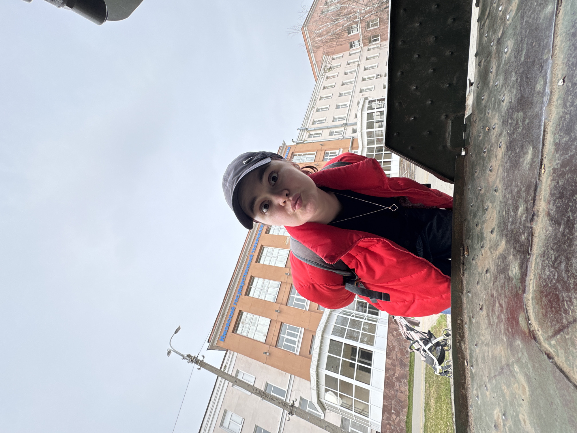 A young person in a red jacket and cap is posing for a selfie. The individual is standing near a weathered, metallic structure, potentially part of a monument or vehicle, given its riveted surface and apparent age. In the background, a multi-story building with windows is visible, and a light pole is positioned to the left. The weather appears overcast, with a grey sky overhead. The location is Izhevsk, Russia, as can be deduced from the building's architecture and potential contextual clues not explicitly visible in the image. A stroller is partially visible in the middle ground between the person and building.