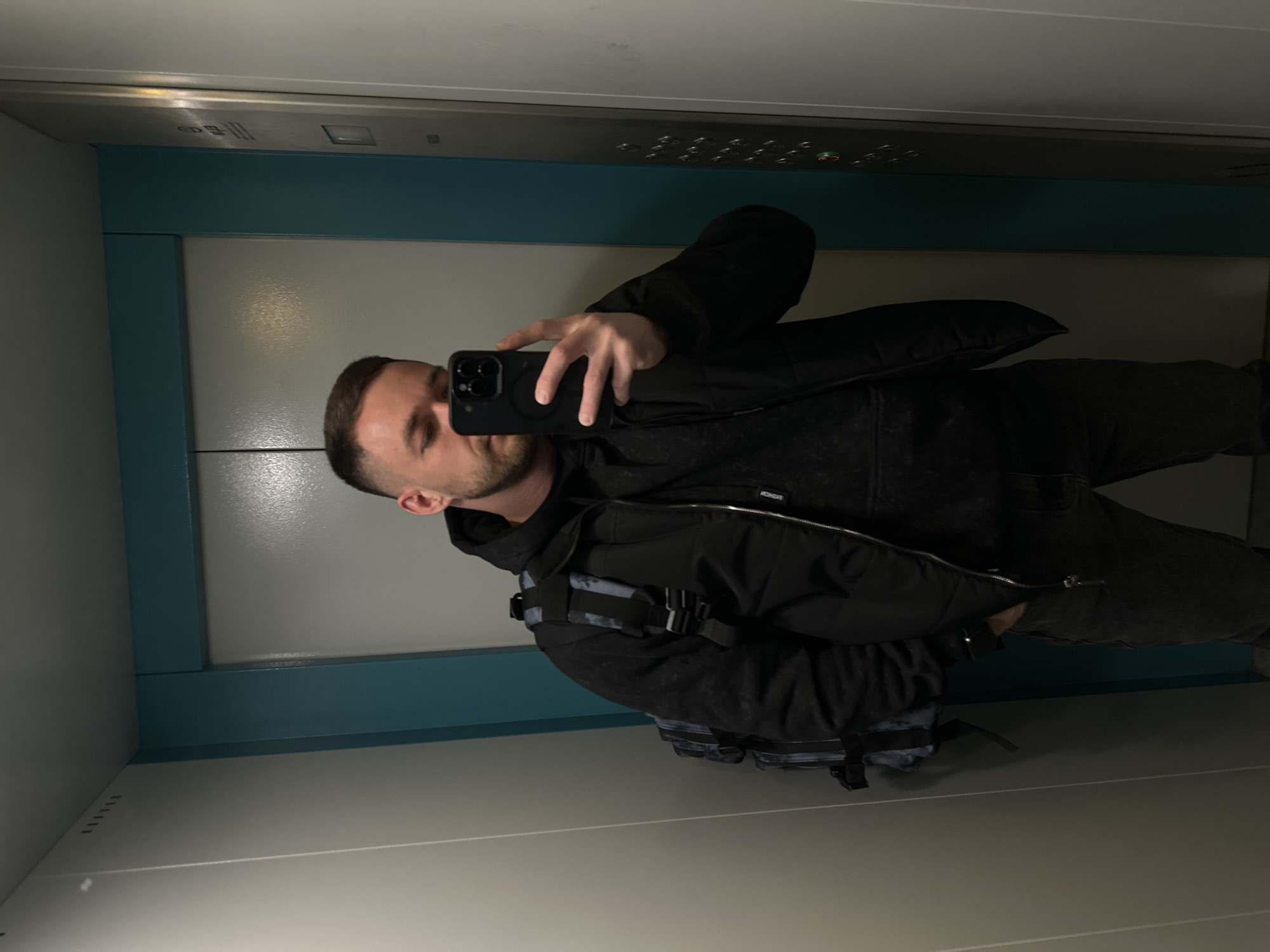 A man is taking a selfie in what appears to be an elevator. The walls of the elevator are light gray with a teal accent stripe near the top. There are elevator buttons above the man. The man is wearing a dark jacket and backpack. The image was possibly taken in the city of Perm, Russia.