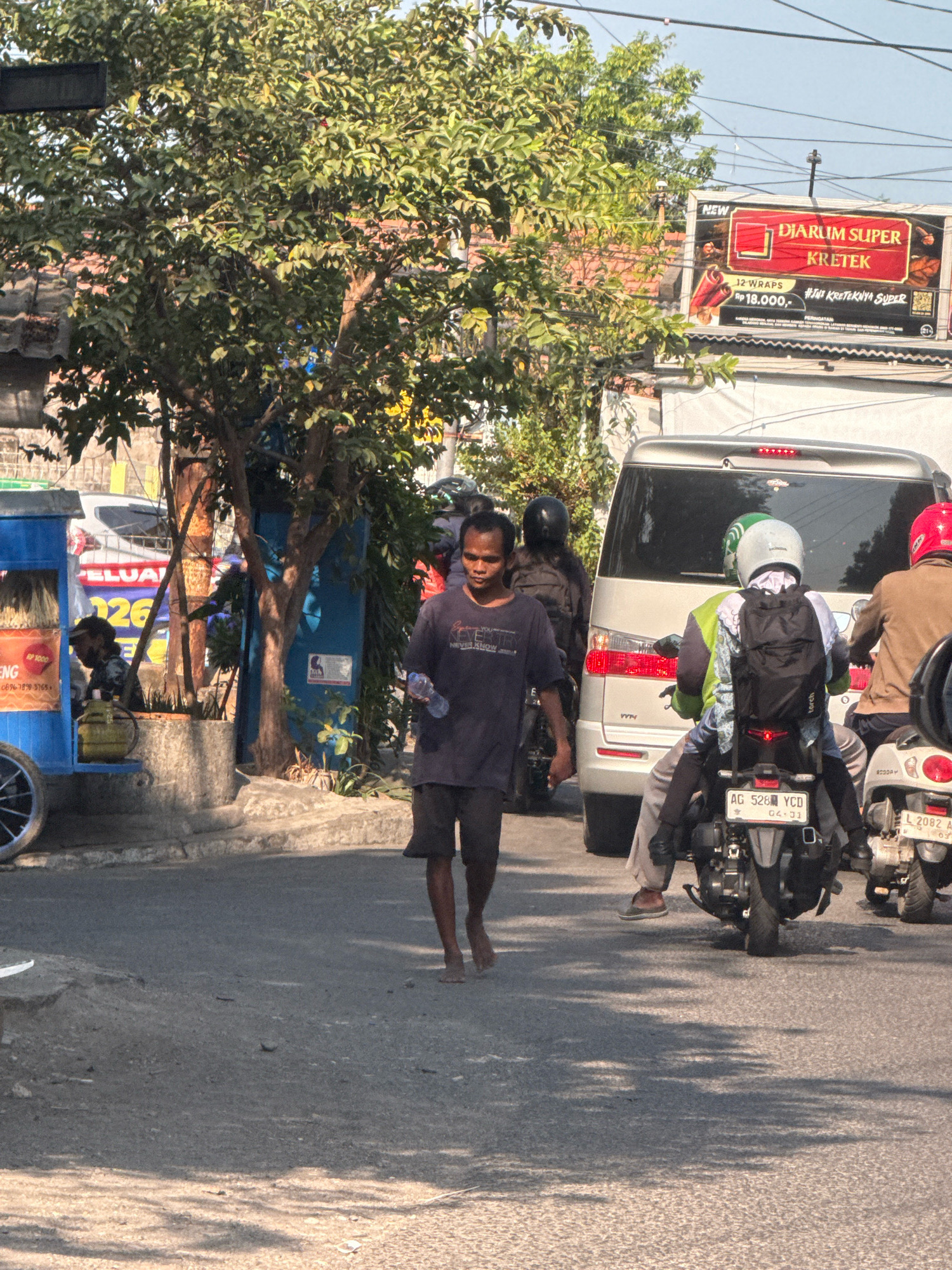 A man walks barefoot on a street in Surabaya, Indonesia, holding a plastic bottle. He is wearing a dark t-shirt and dark shorts. Behind him, several motorcycles are stopped in traffic, including one with a rider wearing a green and white helmet and another with a rider in a red helmet. A silver van is also stopped in traffic. To the left of the frame, a blue food cart with yellow accents is parked. A billboard advertising "DIARIUM SUPER KRETEK" is visible in the background, along with lush green trees and overhead wires. The street appears to be paved, with some cracks and debris. The sun is shining, casting shadows on the ground.
