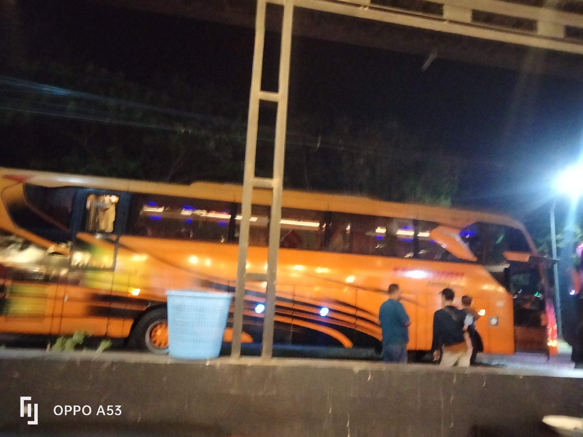 The image shows an orange bus parked at night in what appears to be a public area in Buduan, Indonesia. There are three people standing near the rear of the bus. One person in a blue shirt and jeans stands facing away from the camera. To their right, another person in a dark jacket and light-colored pants is also facing away. A third person, partially obscured, stands next to them. A blue plastic trash can is located to the left of the bus. The bus has reflective decals and illuminated windows. The environment is dimly lit, with streetlights or other artificial illumination present, casting a bright glow on the right side of the image. The background shows trees and power lines. A metal pole is visible in the foreground, obstructing a portion of the bus. The image is slightly blurry and appears to be taken with a low-light camera setting. The bottom left corner displays the text "OPPO A53".