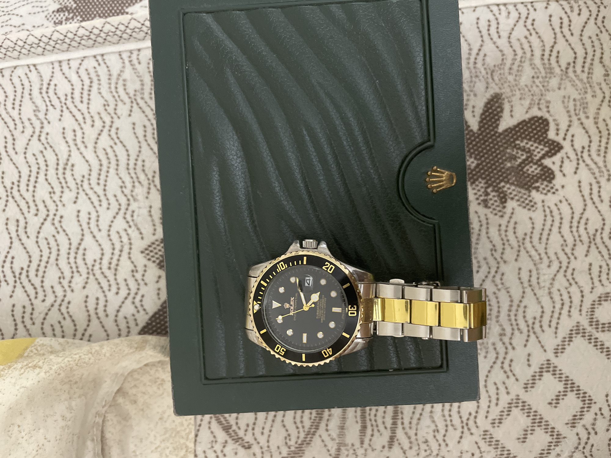 A two-tone watch and its dark green box are prominently displayed, resting on a patterned fabric surface. The watch, styled like a Rolex Submariner, features a black dial with circular and triangular hour markers, and a date window with a cyclops magnifier at the 3 o'clock position. Its bezel is gold with a black insert, marked with dive timings. The watch bracelet is a mix of silver and gold-toned links.
Text visible on the watch dial includes "ROLEX", "OYSTER PERPETUAL", "SUBMARINER", and "SUPERLATIVE CHRONOMETER OFFICIALLY CERTIFIED". The watch box is a deep forest green, with a distinctive wavy or textured pattern on its lid. A golden Rolex crown logo is embossed on the front edge of the box.
The objects are placed on a white or off-white fabric, possibly a bedspread or tablecloth, which is adorned with a repetitive pattern of wavy, dotted brown lines and larger, more scattered brown floral or leaf-like motifs. The scene appears to be indoors, under ambient lighting, captured from a slightly elevated, overhead perspective. No human activity is depicted. The provided location context of Ihtiman, Bulgaria is not visually discernible from the image.