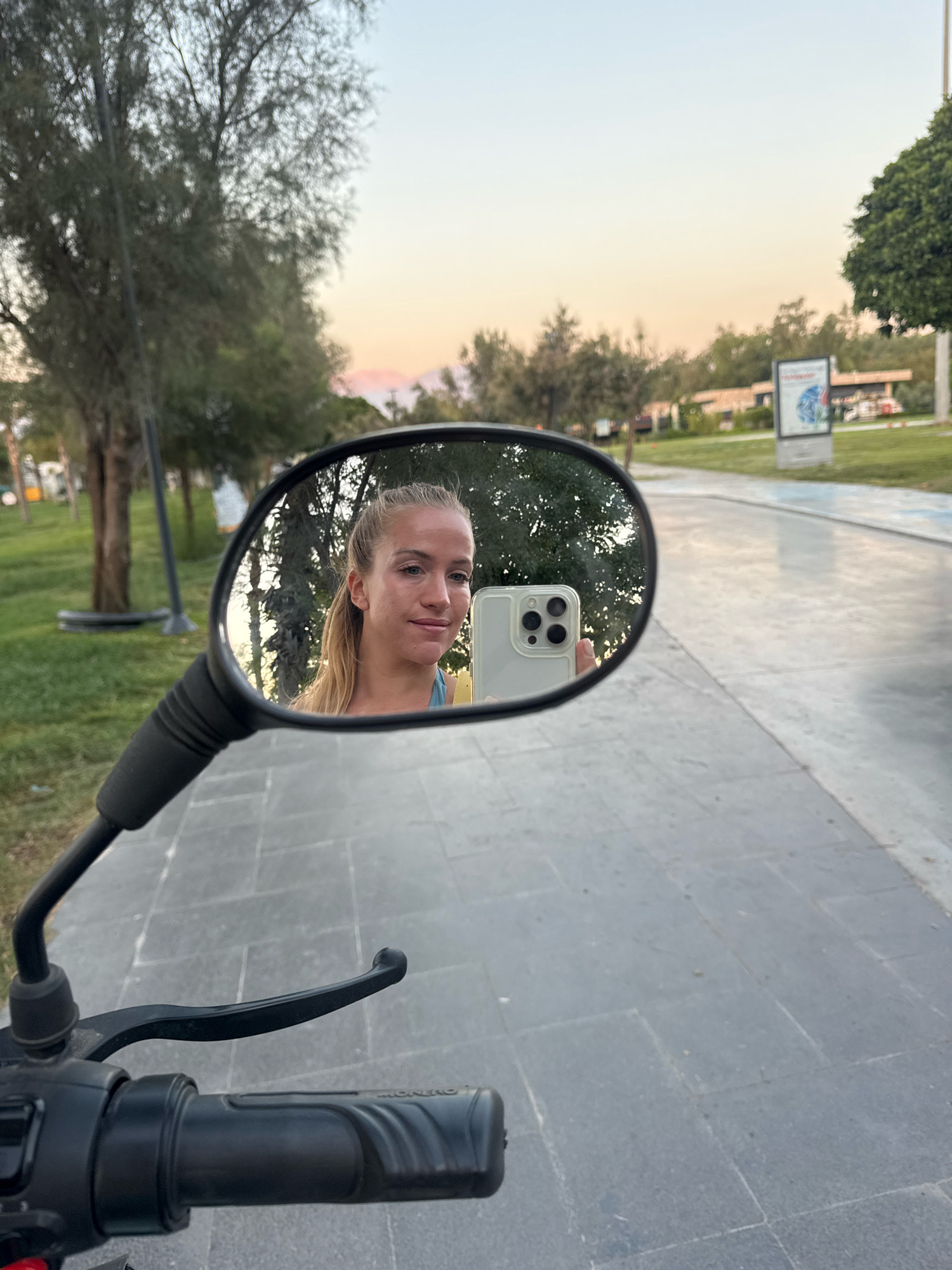 A blonde woman is reflected in a motorcycle mirror, holding a white smartphone to take a selfie. She is wearing a blue top. The background shows a pathway, trees, and a light pole. A billboard is visible in the distance on the right. The scene appears to be outdoors in Antalya, Turkey, possibly near a park or promenade, during a sunset with a pale orange and blue sky. The motorcycle handlebars are in the foreground.