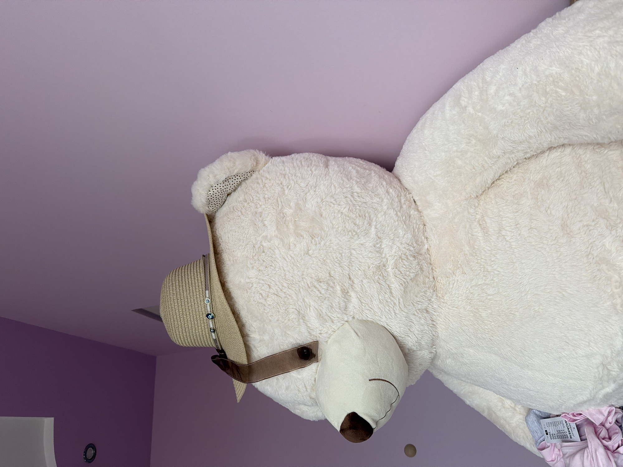 A large, cream-colored plush teddy bear is positioned against a light purple wall in Etropole, Bulgaria. The bear wears a woven straw hat adorned with a gray ribbon featuring three blue and white bead-like ornaments. A brown ribbon strap hangs from the hat over the bear's left eye area, secured by a single brown button. The bear's left ear displays a white fabric lining with black polka dots. Its facial features include a dark brown nose and a stitched smile. The background consists of a wall painted in shades of lavender, showing a corner where the hue shifts. The white ceiling is visible in the upper left, containing a circular recessed light fixture. A small, round, light brown object is affixed to the wall to the left of the bear's head. In the lower left foreground, a small pile of fabric, including pink ruffled material and a partially visible clothing tag reading "DEPOCO WOMEN," rests beside the bear.