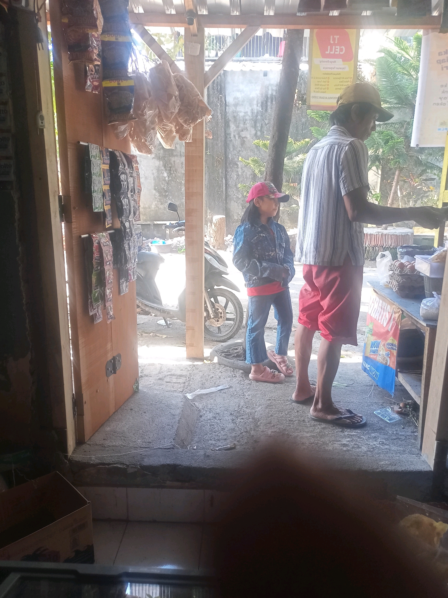 In a street-side kiosk in Praya, Indonesia, a man in a striped shirt and red shorts stands behind a counter, facing away from the camera. He appears to be engaged in a transaction. To his left, a young girl wearing a denim jacket, red shirt, blue jeans, and a pink baseball cap stands looking to her left, her right hand near her face. She wears pink sandals. A motorcycle is parked to the left of the girl. The kiosk is partially open, with wooden doors on the left. Bags of snacks hang from the wall inside the kiosk. The ground is a concrete surface. Sunlight streams into the scene from the right.