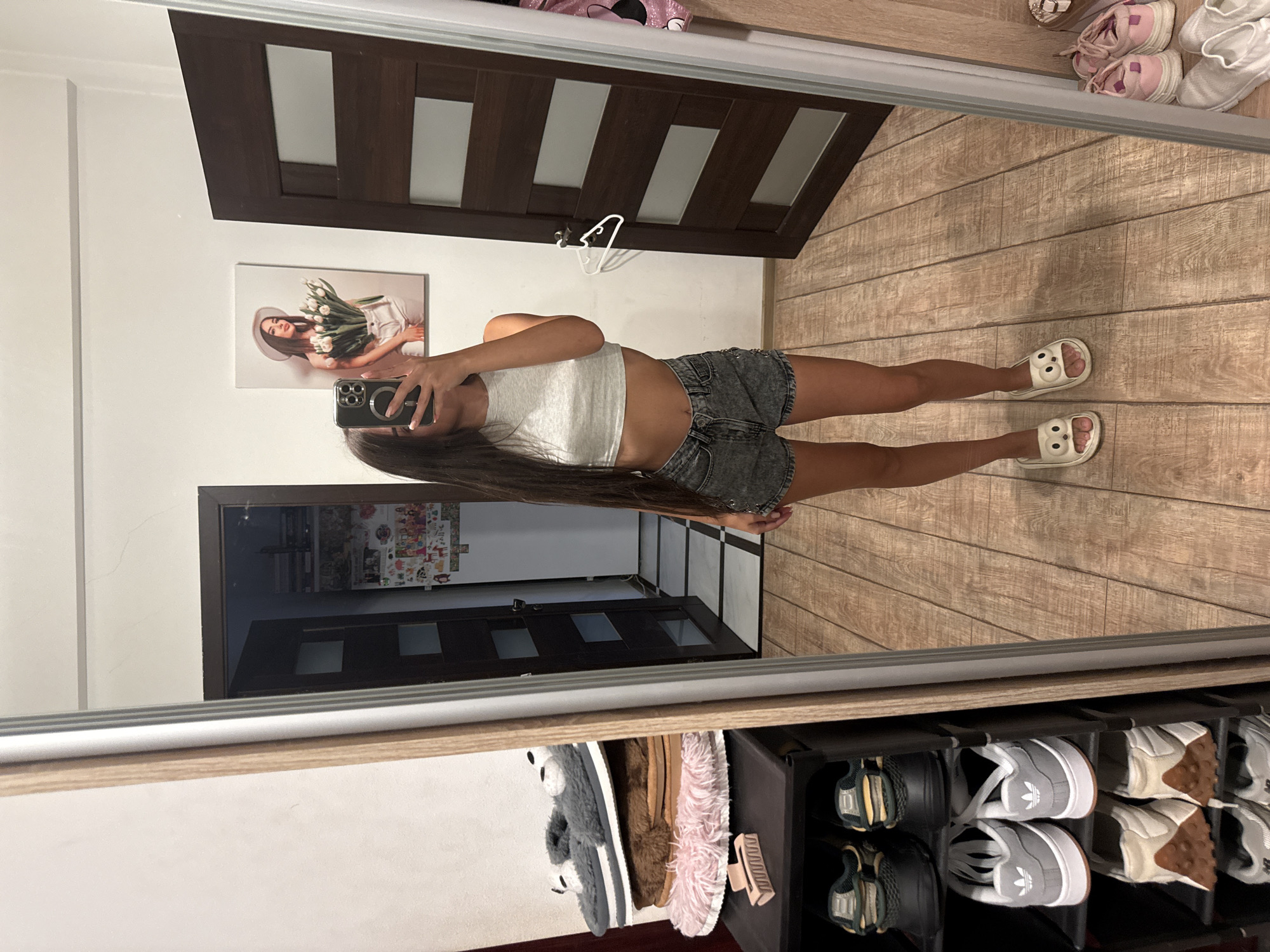 A young woman is standing in front of a full-length mirror in what appears to be a residential setting, possibly an apartment in Chișinău, Moldova. She is taking a mirror selfie, holding her smartphone in her right hand. She is wearing a grey cropped tank top and denim shorts. Her long, dark hair cascades down her back.
The room is furnished with a shoe rack filled with various pairs of sneakers and slippers. On top of the shoe rack, a stack of fluffy slippers is visible. To the left of the shoe rack, a partially opened dark wooden door leads into another room, where a black shoe organizer is visible in the reflection. On the wall behind her, a framed picture of a woman holding a bouquet of white tulips is displayed. To the right, another dark wooden door with glass panels is visible. The floor is covered with wooden planks. The overall impression is that of a casual, personal space within a home.