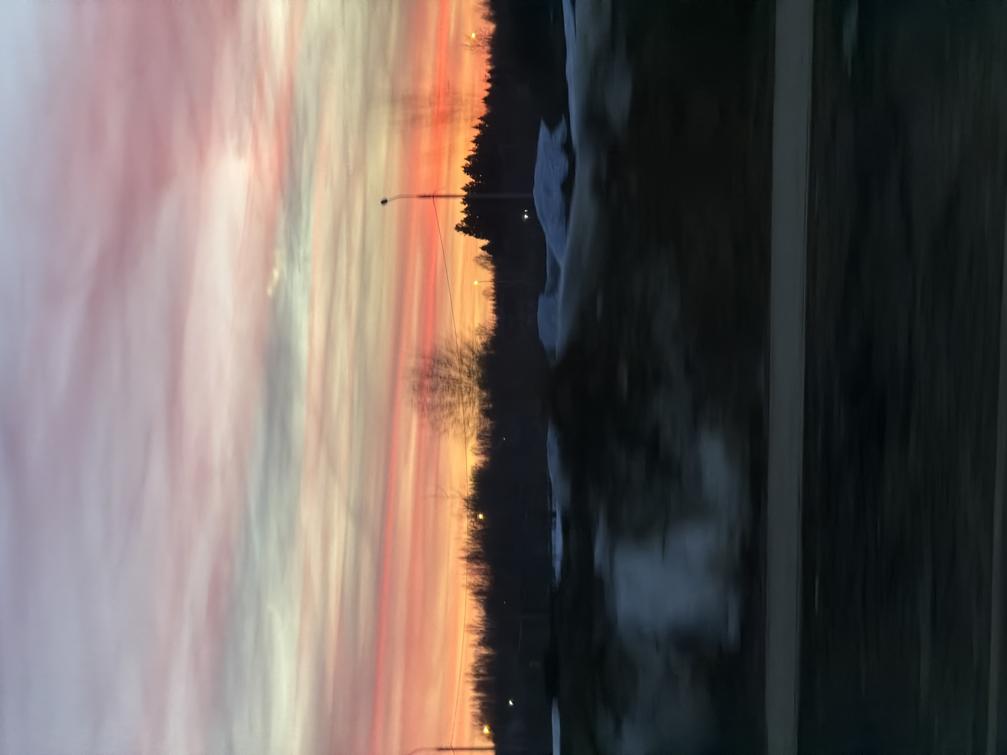 The image shows a landscape at dusk or dawn. To the left, a colorful sky transitions from pink to orange. A treeline divides the sky from the ground. Streetlights are visible along the treeline. On the right, a dark, indistinct area possibly depicts snow and infrastructure. Given the specified location of Berezniki, Russia, the setting likely depicts a winter scene. No people are visible.