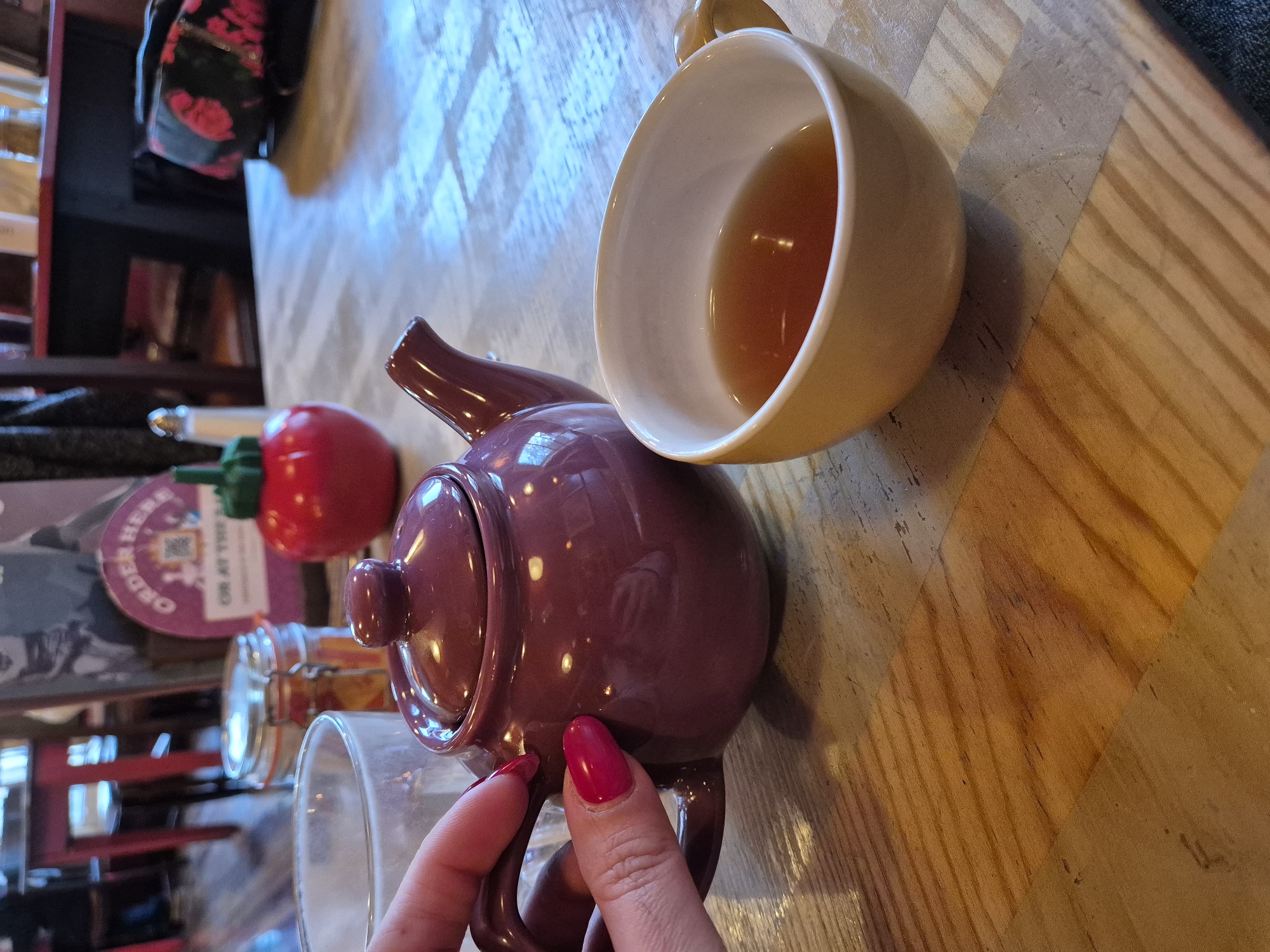 The image depicts a tea service on a wooden table. A partially filled yellow teacup rests on the table beside a dark purple teapot. A hand with red nail polish is holding the teapot's handle. Other objects are in the background, including a red decorative object, a glass jar with contents, and a partially visible glass. The scene is likely located in West Suffolk, United Kingdom, based on the text on the image, which is not clearly visible.