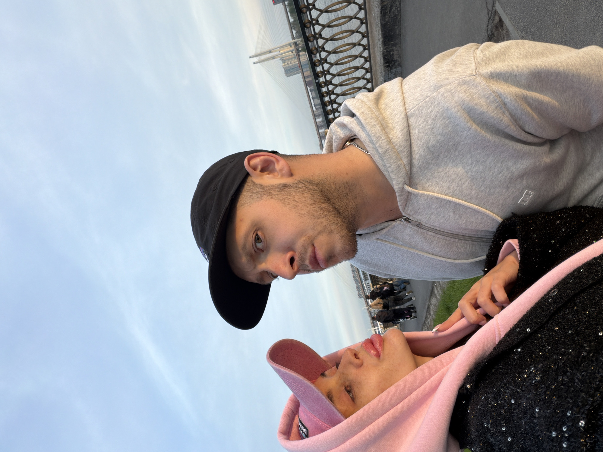 A man and a woman are posing for a photograph outdoors. The man is wearing a black baseball cap and a gray hooded sweatshirt. The woman is wearing a pink hooded top with a black, sparkly jacket. The setting appears to be Saint Petersburg, Russia. A cable-stayed bridge is visible in the background, along with an ornamental metal fence.