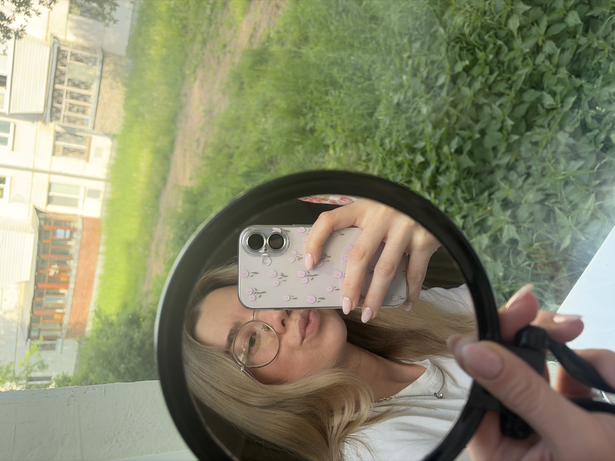 A woman takes a selfie in a round mirror, partially covering her face with her phone. The phone has a clear case with small pink floral patterns. The woman has blonde hair, wears round glasses, and a white shirt with a silver necklace. Her fingernails are polished with a light pink color. Reflected in the mirror are buildings, green grass, and trees. The location is Chaikovsky, Russia.