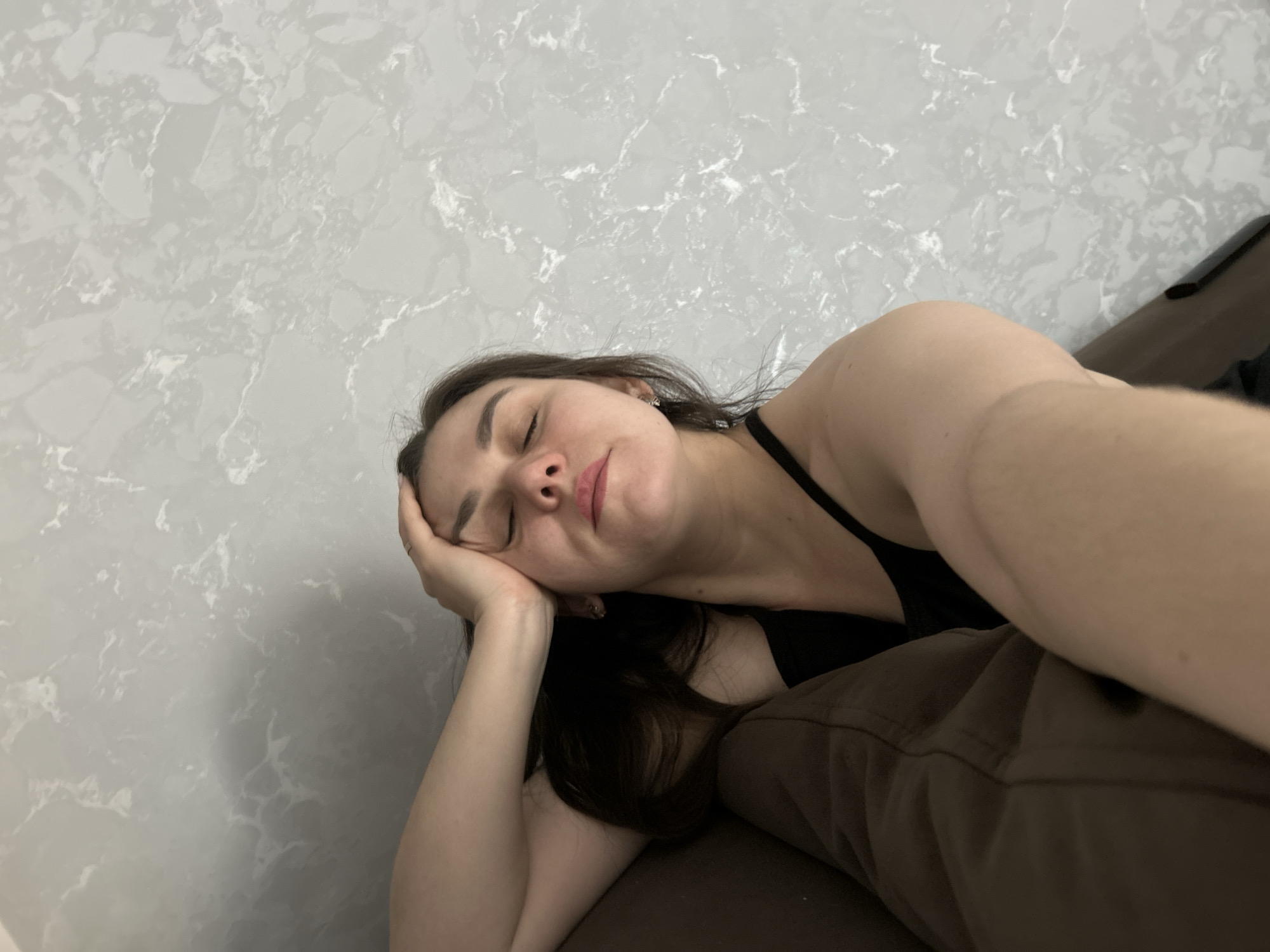 A young woman with dark hair and fair skin is lying down, her head resting on her hand. She appears to be sleeping or resting with her eyes closed. She is wearing a black tank top and brown pants. The background is a wall with a gray and white marble pattern. The scene is indoors. There is no visible information to confirm the location as Chaikovsky, Russia.