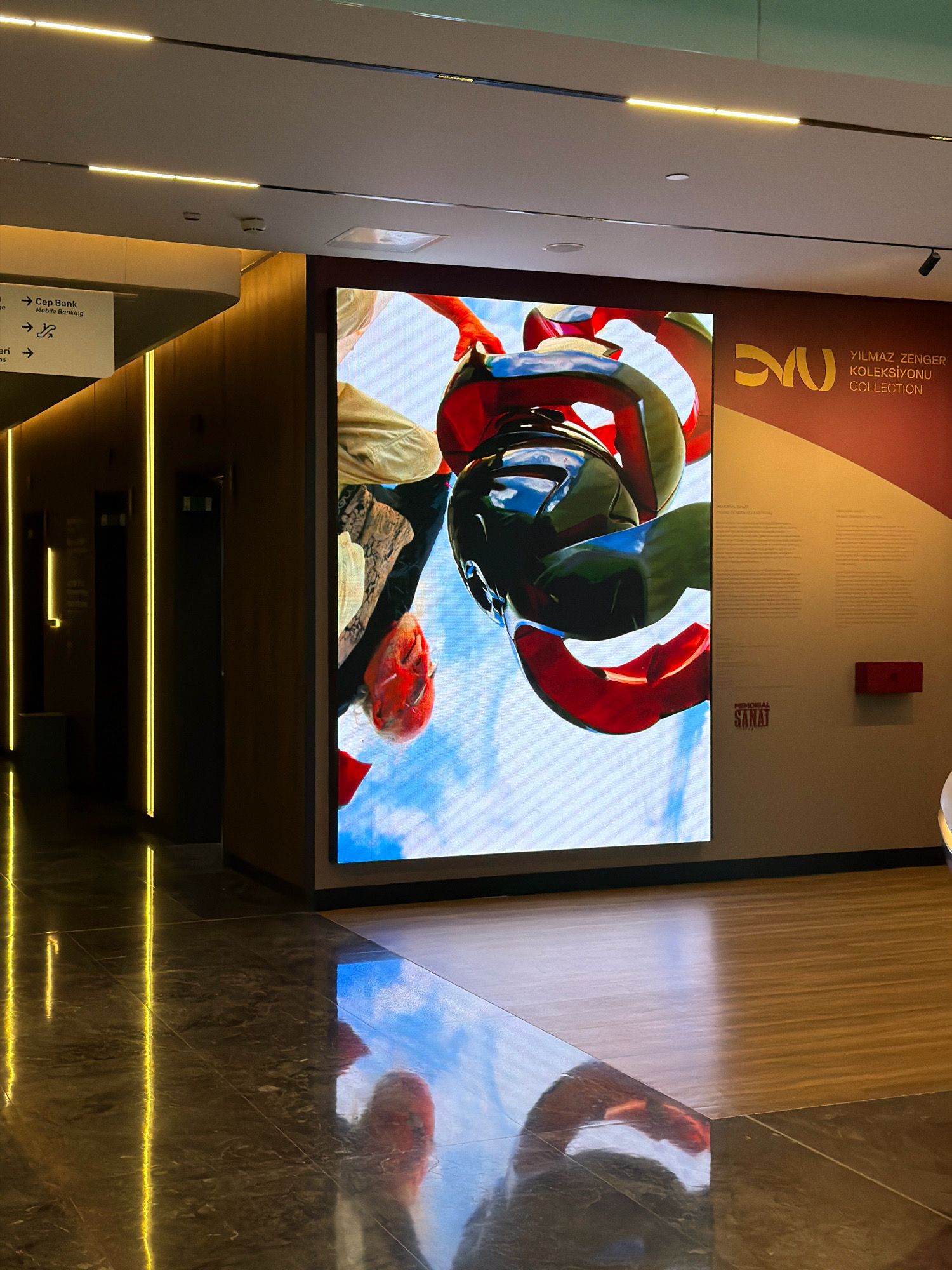 The image depicts an indoor hallway with a large digital screen displaying a close-up of a person's head, possibly adorned with a helmet, against a sky backdrop. To the left, a directional sign points towards "Cep Bank" and "Mobile Banking." The hallway features modern lighting with yellow LED strips on the walls and ceiling, and a polished marble floor reflecting the lights and the digital screen. On the right wall, there is an exhibition display with text and a logo that reads "YILMAZ ZENER KOLEKSIYONU COLLECTION" and "SANAT." This scene appears to be located in Ataşehir, Turkey, likely within a cultural institution or a modern building complex.