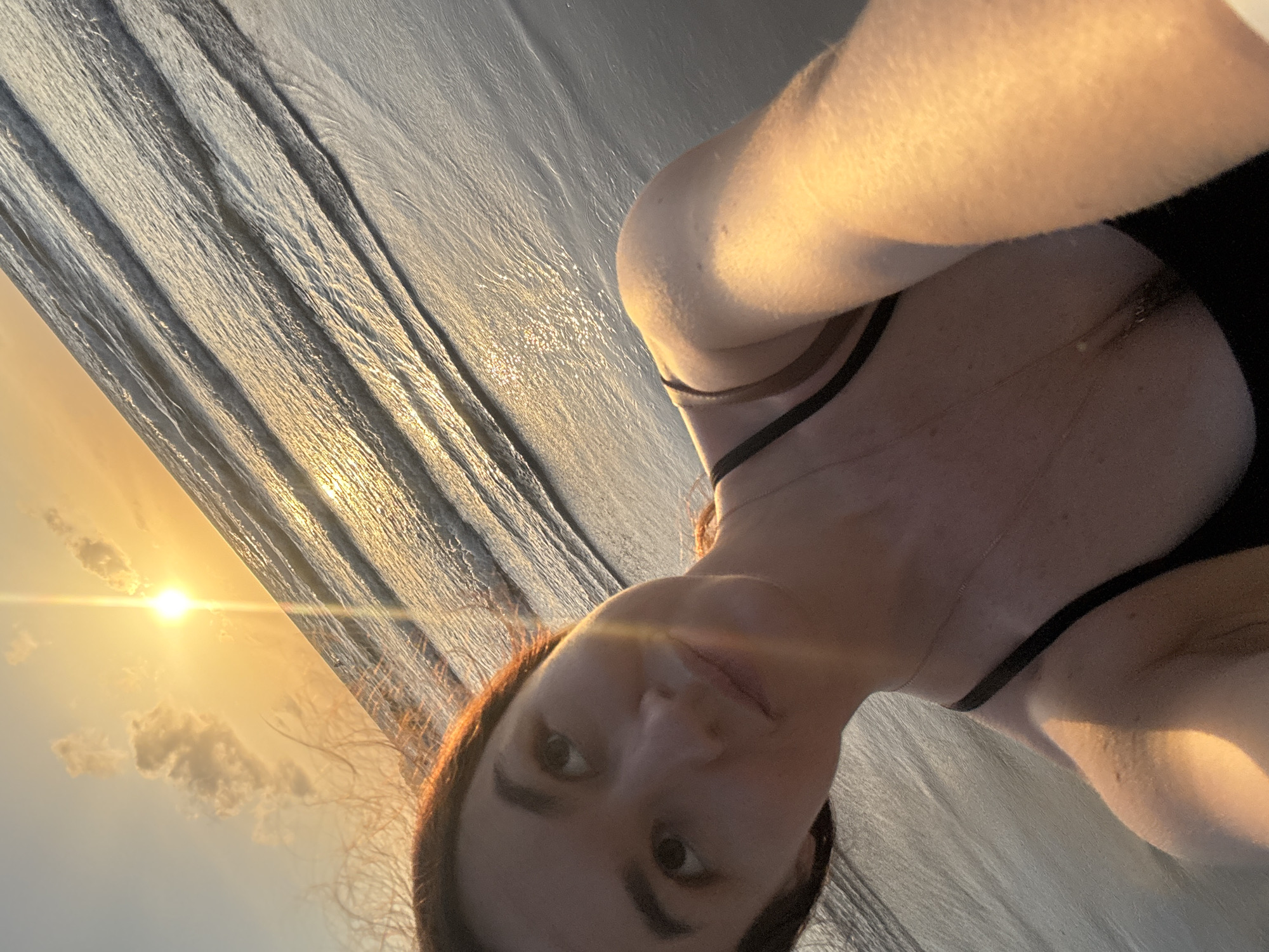 The image shows a young woman taking a selfie on a beach at sunset. She is wearing a black swimsuit and a gold necklace. The sun is setting over the ocean, casting a warm glow on the water and the woman's face. The waves are gentle, and the sand is wet. The woman appears to be enjoying the sunset and the peaceful atmosphere of the beach. The location is Patong Beach in Phuket, Thailand.