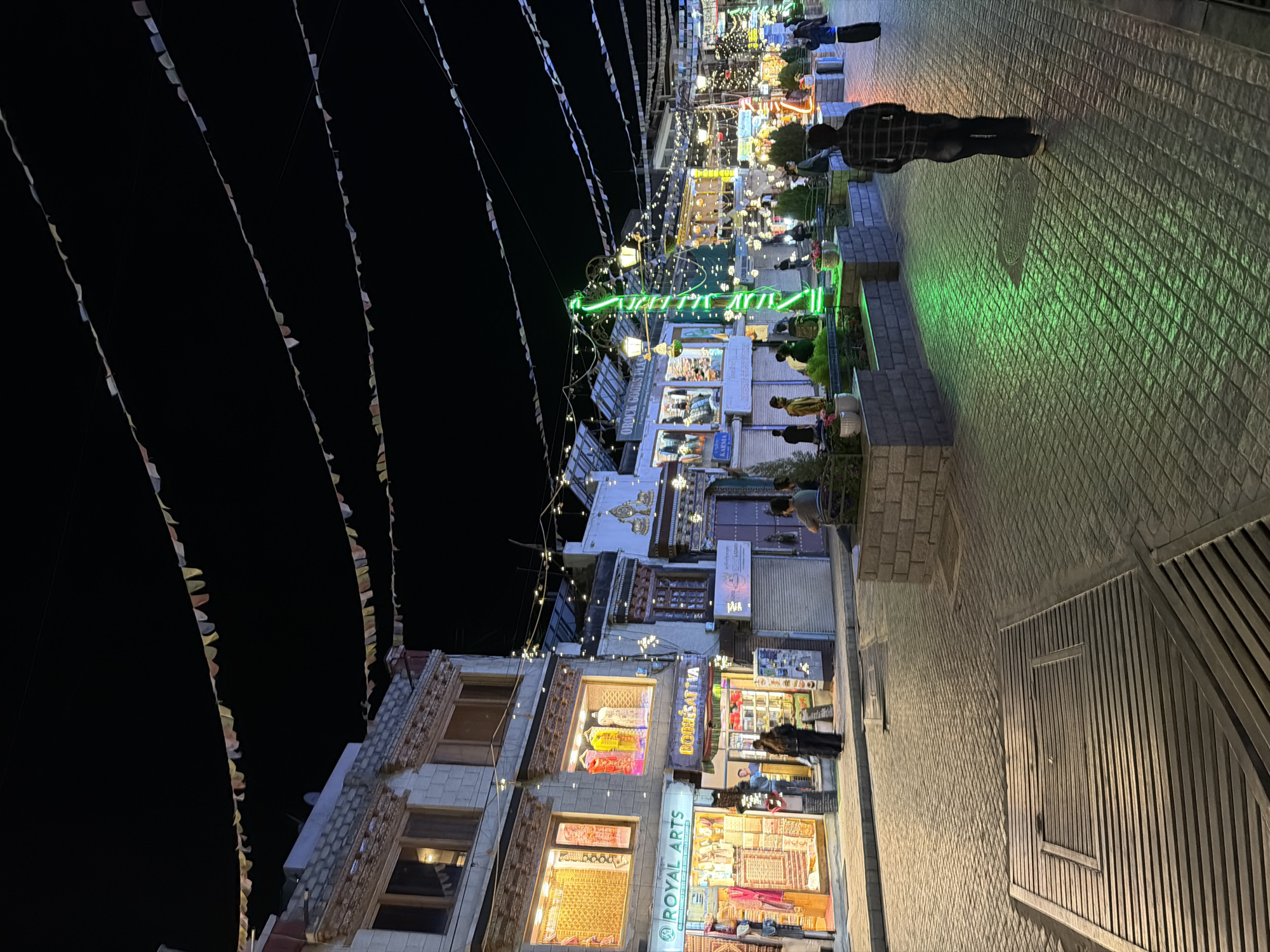 This is a nighttime photograph of a street in Leh, India. The street is paved with cobblestones, and there are shops lining both sides. Prayer flags are strung across the street overhead. The shops are illuminated, and some have displays of clothing and other goods. There are several people walking on the street, and some are sitting on benches. A man in a plaid shirt and jeans is walking away from the camera, and a woman in a black dress is standing in front of a shop. There is a significant amount of green lighting emanating from a display in the middle of the street, likely from decorative lights. The overall scene depicts a lively street scene in Leh at night.