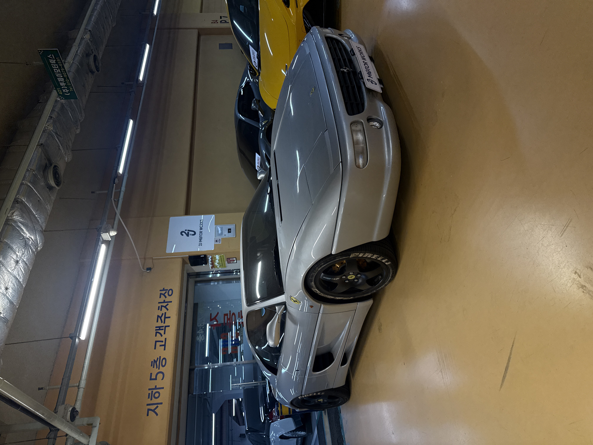 The image depicts an indoor parking garage, specifically "B5 Customer Parking," likely located in the city of Seoul, South Korea. The foreground is dominated by a silver Ferrari with "2J MOTOR WORKS" detail. The image is taken in a somewhat narrow space with at least two other cars parked behind the Ferrari, including a yellow vehicle. Overhead fluorescent lights are visible, as well as exposed ductwork. Signs for "2J MOTOR WORKS," are seen on the wall.