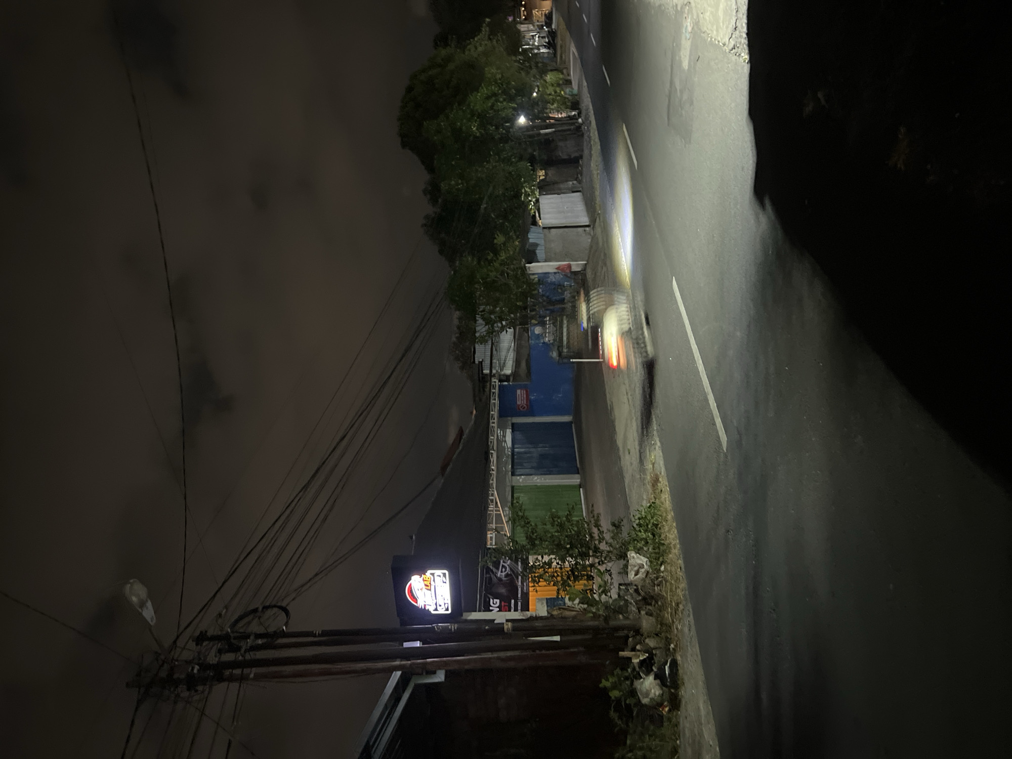 A night scene on a road in Denpasar, Indonesia, shows a dark sky with scattered clouds and a street with a white line marking the lanes. A vehicle's headlights illuminate the road and the foreground. On the left side of the road, a building with a brightly lit sign that reads "Speed Lab" is visible. Utility poles with numerous wires are also present. The building has different colored doors, including yellow, green, and blue. Trees and foliage line the side of the road, and there is some debris on the ground near the building. The scene appears to be in an urban or suburban area, with the lights suggesting commercial activity.