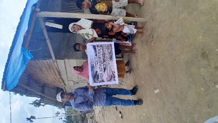 People stand in front of a makeshift house. They hold a sign of appreciation that reads "Thank you Prof. Kathy Berry Penot for your generous donation." Adults, children, and a baby are visible. The house has a blue tarp roof and corrugated metal siding. The ground is dirt and sparse vegetation is visible in the background.
