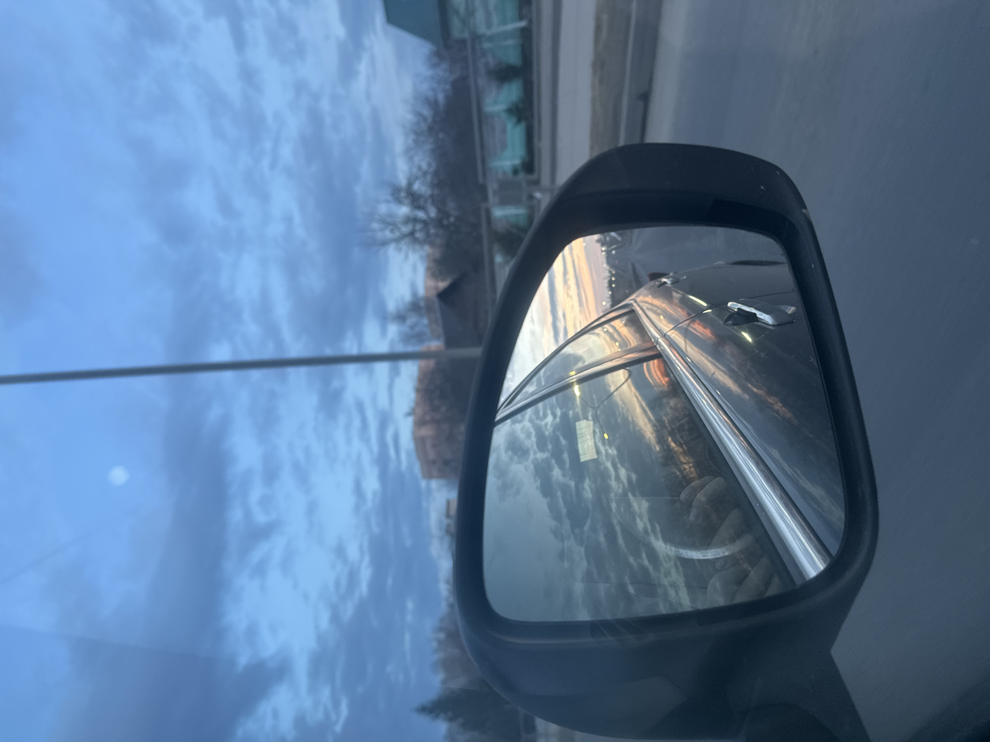 The image, captured from within a vehicle in Liozna, Belarus, presents a view dominated by the car's left side mirror in the lower-left foreground. The mirror reflects the rear section of the vehicle itself, including a chrome window trim, a door handle, and a blurred hand, presumably the driver's, gripping a steering wheel. The reflection also shows a road extending into the distance, with multiple bright points of light suggesting streetlights, and a sky exhibiting orange and pink hues indicative of a sunset or sunrise. Outside the vehicle, the upper portion of the frame is filled with a cloudy, blue-grey sky featuring a distinct, bright circular object, possibly the moon or a luminous light source, in the upper-center. A slender, dark vertical utility pole is visible extending upwards near the center of the frame. The background landscape includes bare deciduous trees on both sides. On the left, a multi-story building with a light-colored facade is partially visible. On the right, a building with a dark, peaked roof and a lighter facade stands behind a fence or barrier featuring transparent or reflective panels. The car appears to be on a paved road, a section of which is visible in the lower-right corner of the image.