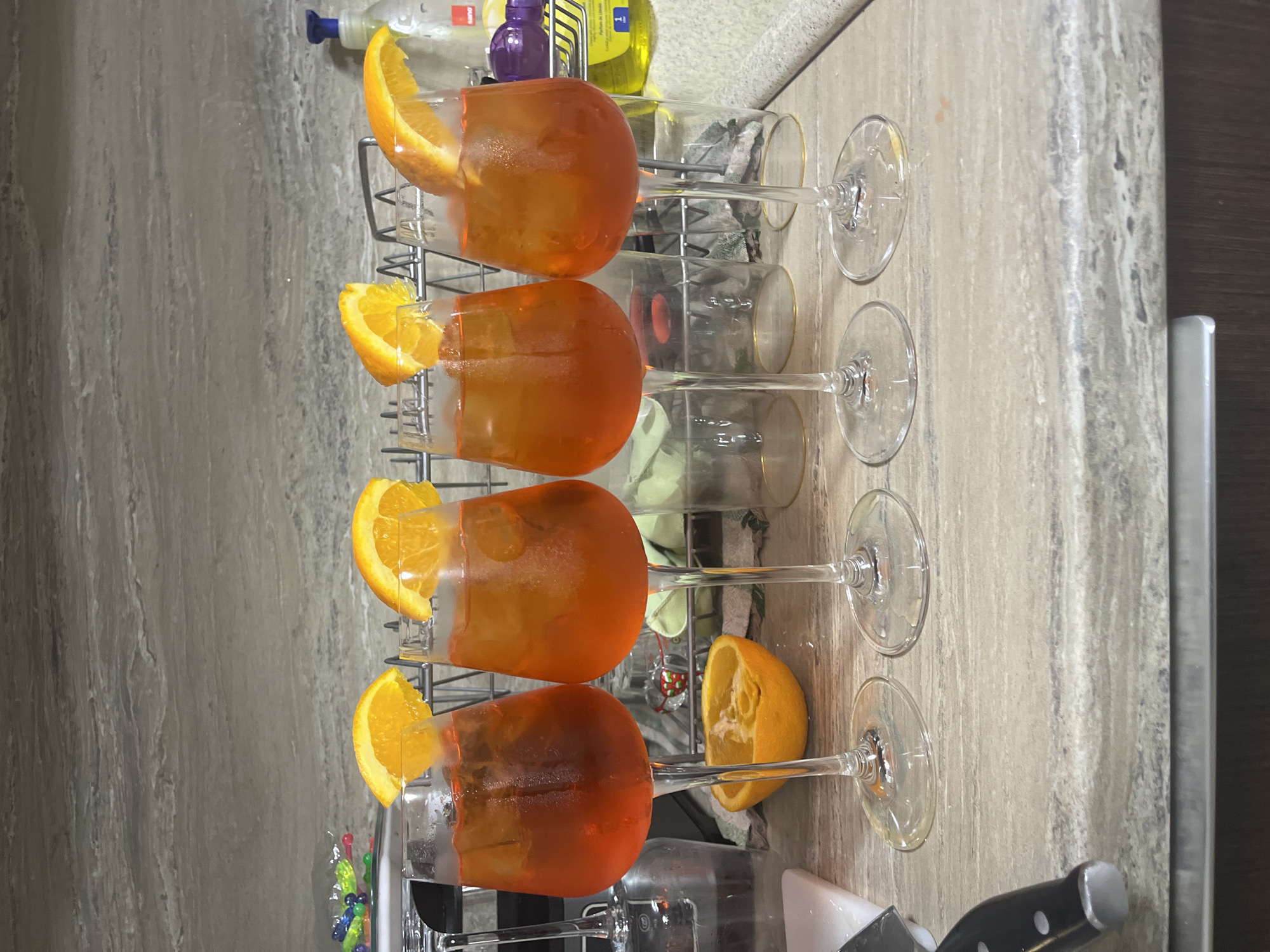 The image shows four filled wine glasses, likely containing an orange-colored beverage. Each glass is garnished with a slice of orange on the rim. The glasses are arranged in a metal dish rack on a kitchen counter, which has a mottled gray and tan surface. Additional kitchen items, including bottles and a knife, are partially visible in the background. There is no information indicating a specific location within Asenovgrad, Bulgaria.