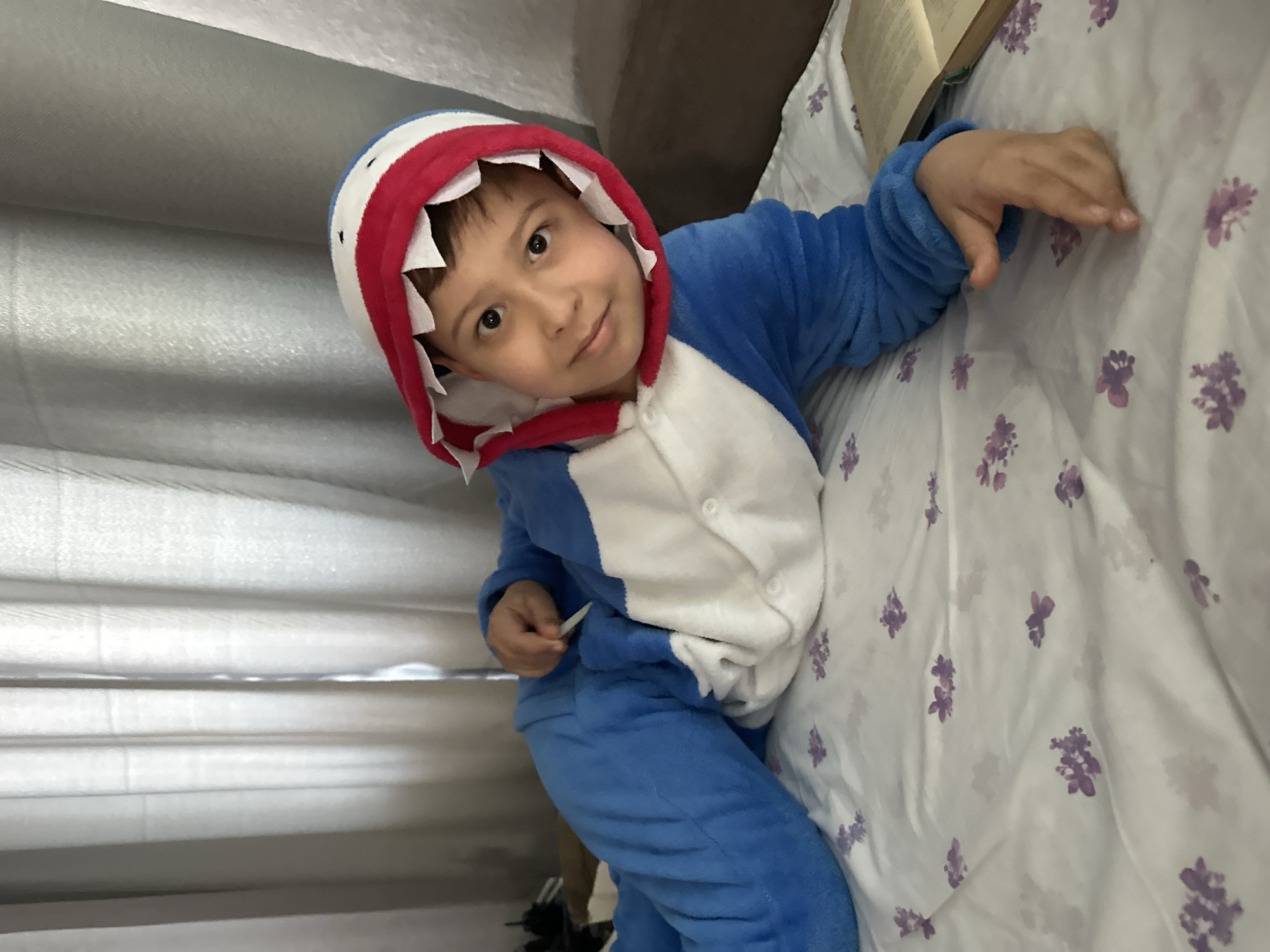 A young child, likely a boy, is depicted indoors, wearing a blue and white shark-themed onesie or kigurumi. The child is positioned on a surface, possibly a bed, covered with a white sheet adorned with scattered purple floral patterns. The image is taken from a slightly elevated perspective and is rotated counter-clockwise, creating a tilted view.
The child, with dark hair and brown eyes, looks directly at the camera with a gentle, engaging smile, conveying a sense of happiness and comfort. His head is framed by the shark hood, which features red trim and white fabric 'teeth' lining the opening. His right arm is extended and rests on the patterned sheet, while his left hand holds a small, thin white object, possibly a piece of paper or a small item.
In the background, a light-colored curtain or pleated blind with horizontal folds is visible, suggesting an indoor setting like a bedroom. A partial view of a book is discernible behind the child's right arm, resting on the bedsheet. No legible text is visible on the book or elsewhere in the image. The lighting is soft and even, typical of an indoor environment, making the time of day indeterminate. The scene captures a playful and relaxed moment.