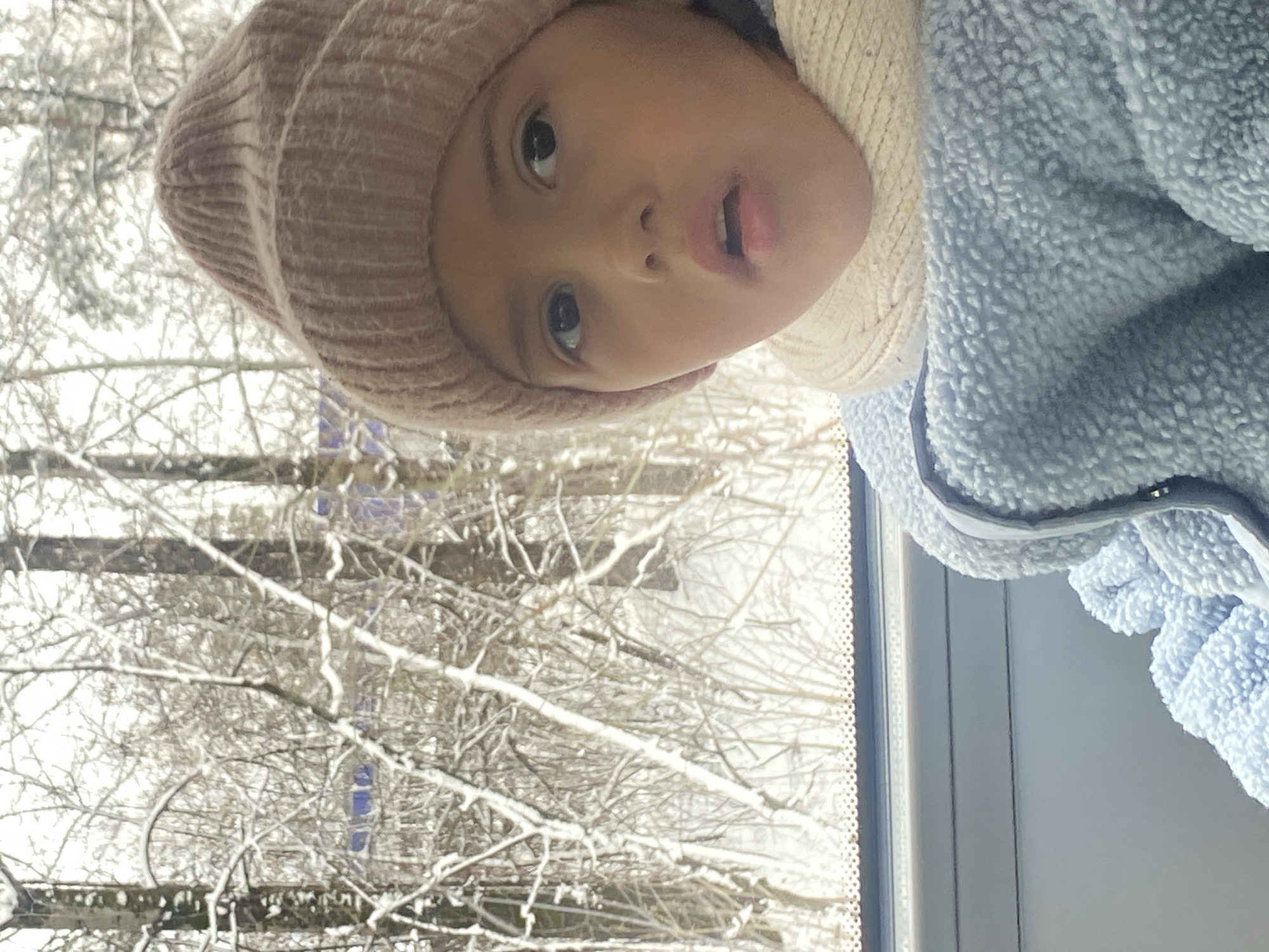 A child wearing a pink knit hat and a white scarf looks up towards the camera, with a blue, fuzzy jacket visible on their right side. Through a window behind them, bare, snow-covered tree branches are visible. The location is Chaikovsky, Russia.